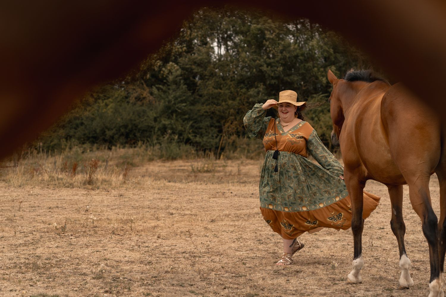 Woman in vintage western dress saluting near a brown horse in a dry field with trees in background.