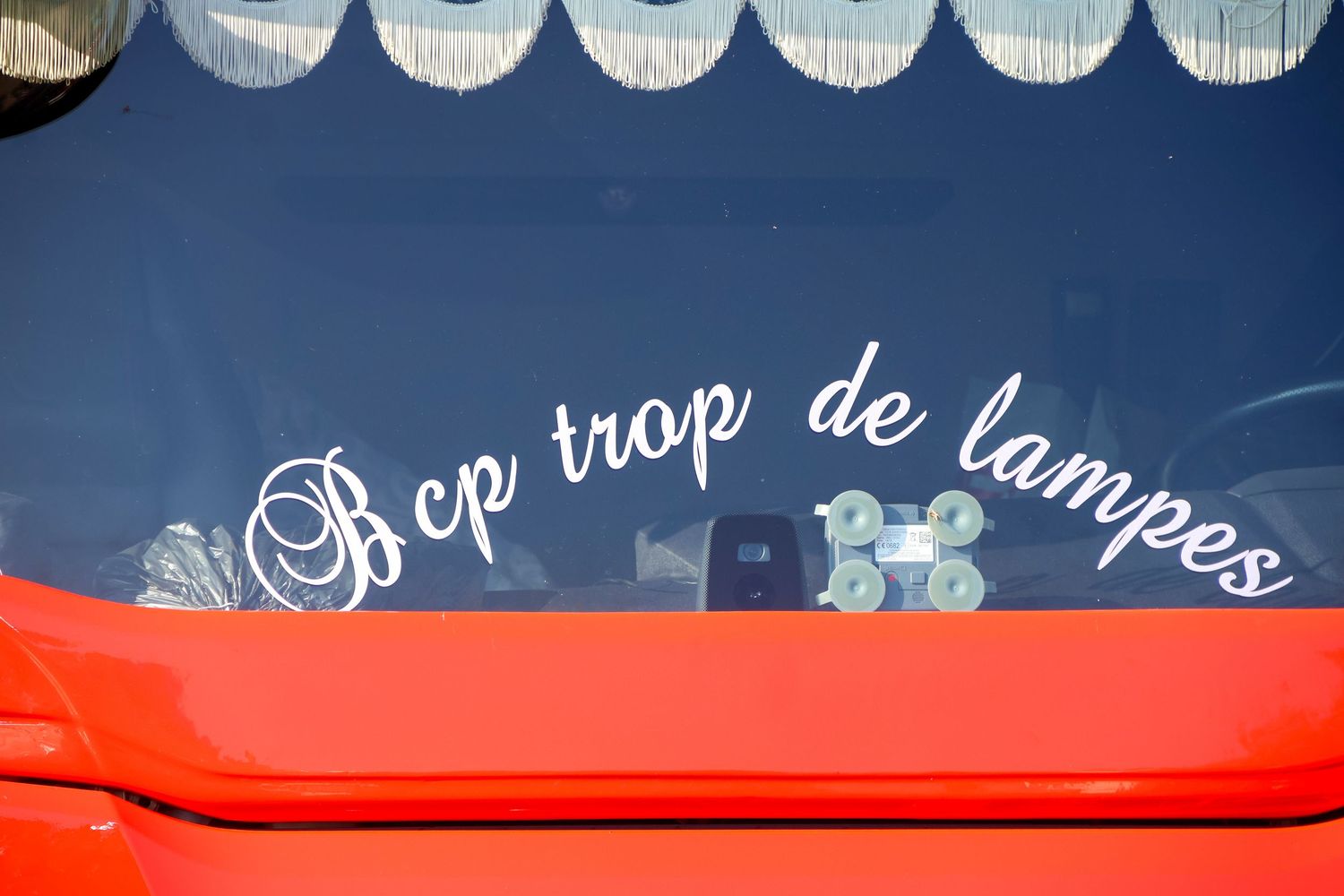 Red truck rear window displaying white cursive text reading Bcp trop de lampes with decorative curtains inside.