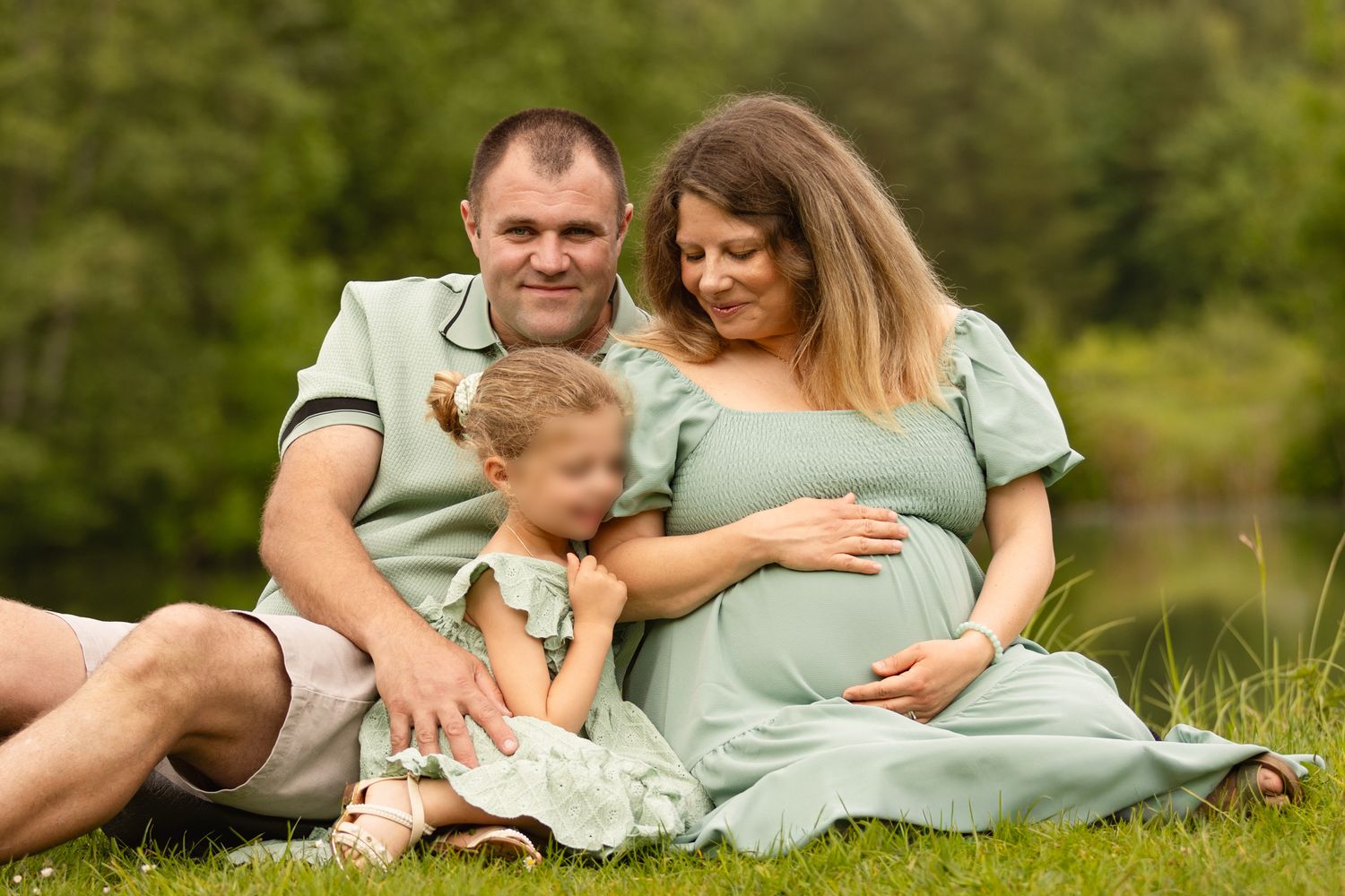Happy pregnant family sitting on green grass outdoors, parents and child in matching sage green outfits.