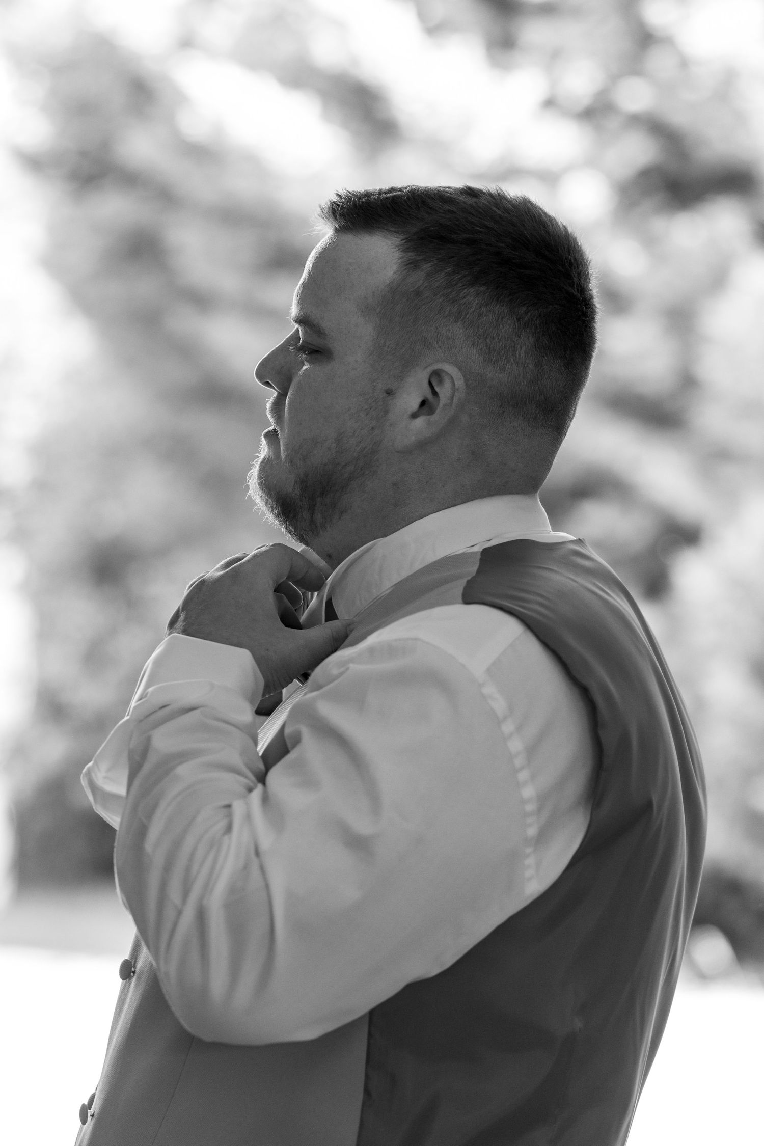 Black and white photo of a groom adjusting his bow tie while wearing a vest outdoors.