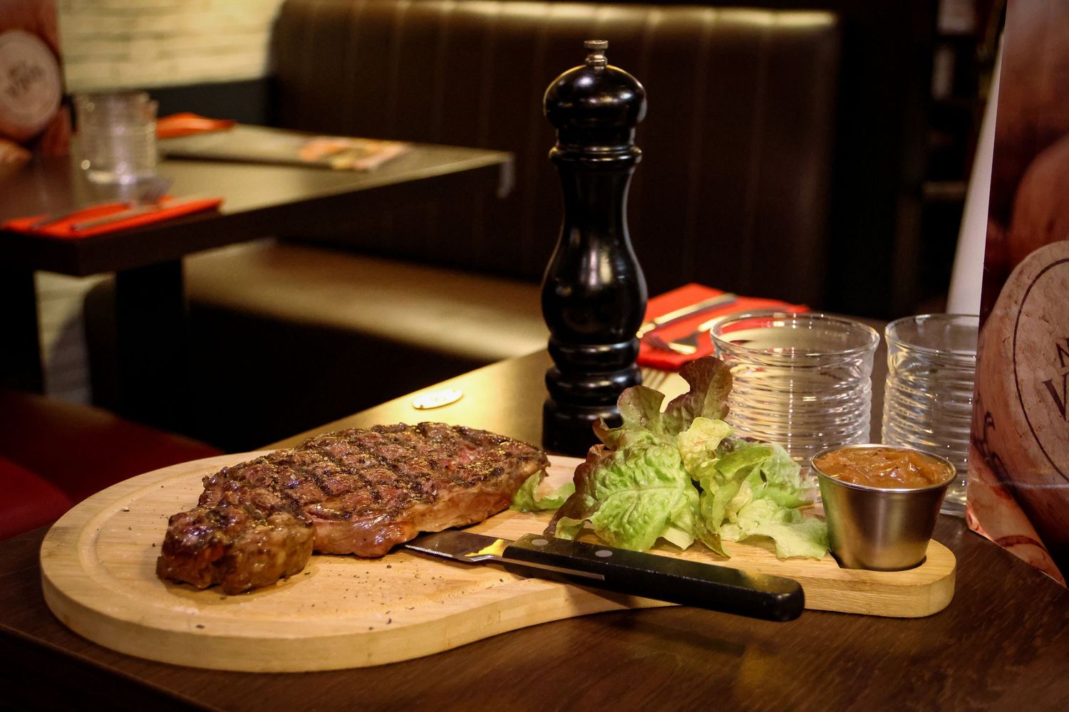 A grilled ribeye steak served on a wooden board with fresh salad and dipping sauce beside a pepper mill.