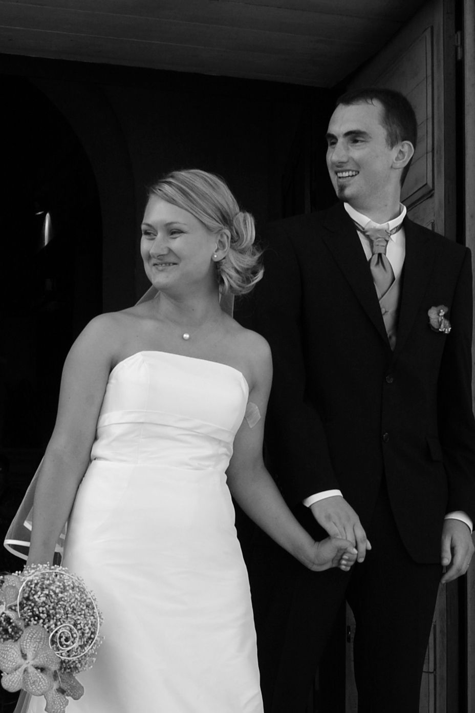 Black and white photo of a smiling bride in strapless gown holding bouquet with groom in suit exiting a church.