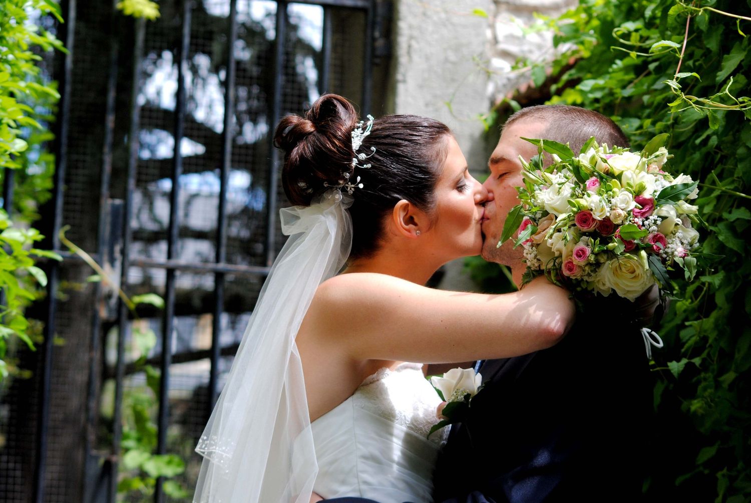 A bride and groom sharing a kiss while holding a floral bouquet at an ornate garden gate.