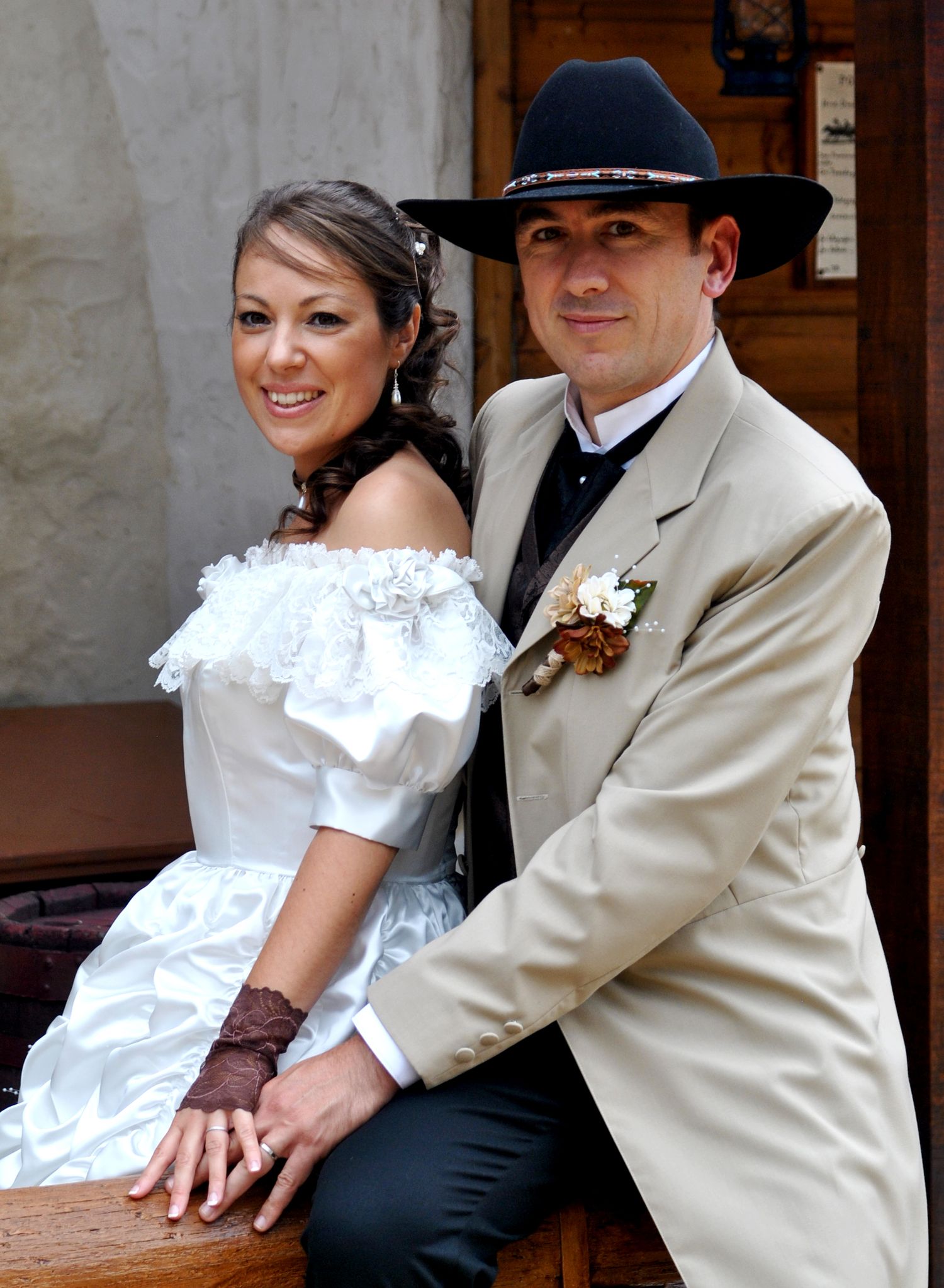 Western wedding couple posing together, bride in white off-shoulder gown and groom in tan suit with black cowboy hat.