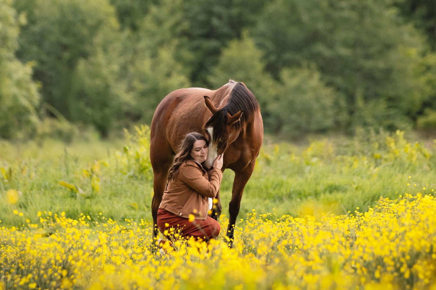 Woman sitting in yellow wildflower field with brown horse nuzzling her in a lush green meadow.