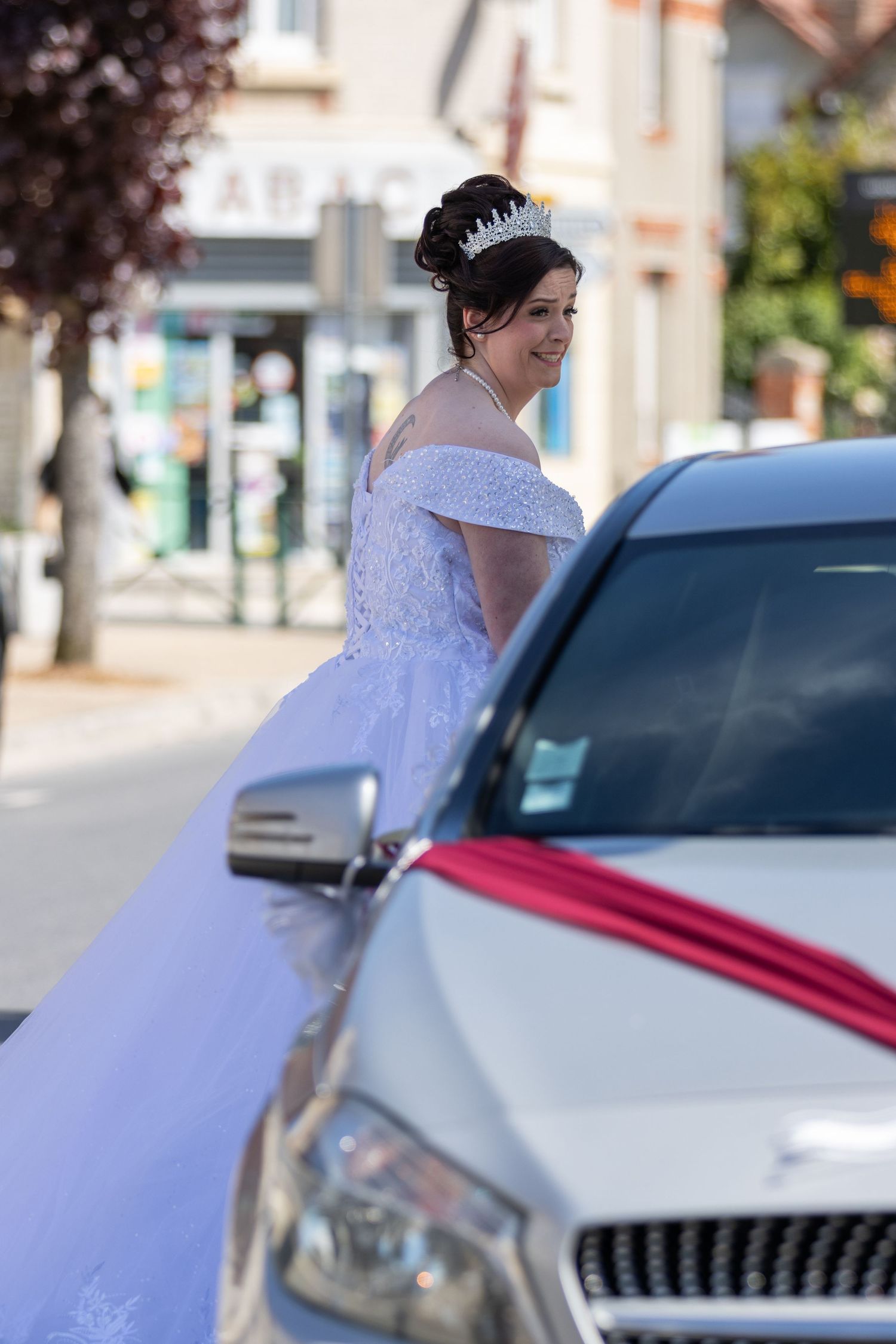 Smiling bride in white off-shoulder gown and tiara poses beside a decorated wedding car on a sunny street.