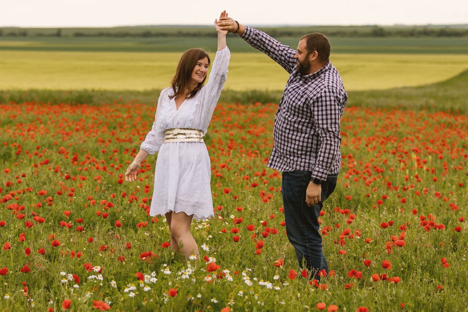A couple dances joyfully in a vibrant red poppy field, the woman in a white dress and man in a plaid shirt.