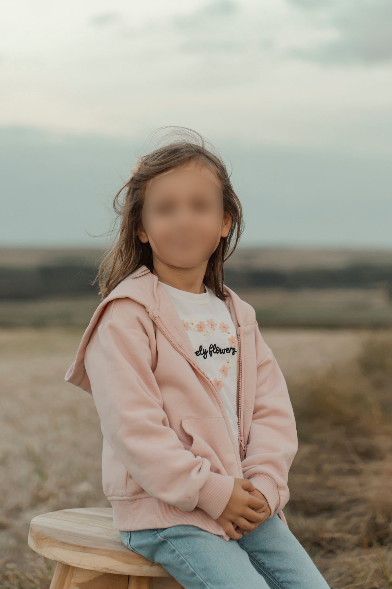 Young girl in pink hoodie and jeans sitting on wooden stool in open field with cloudy sky backdrop.
