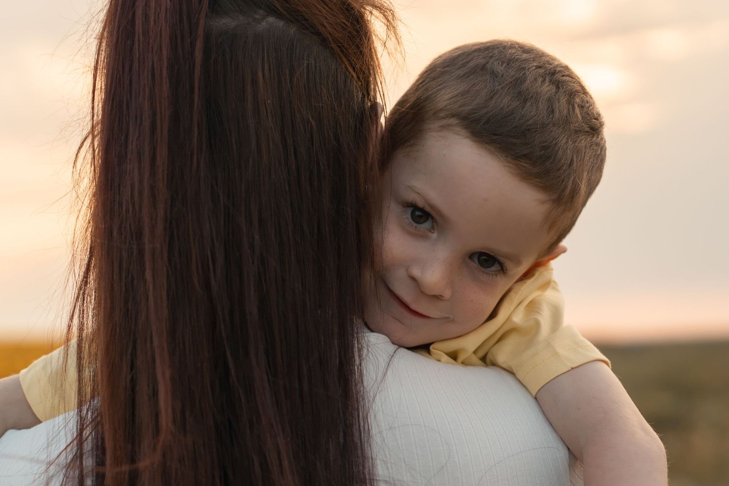 A young child resting their head on a woman's shoulder during a warm sunset outdoors.