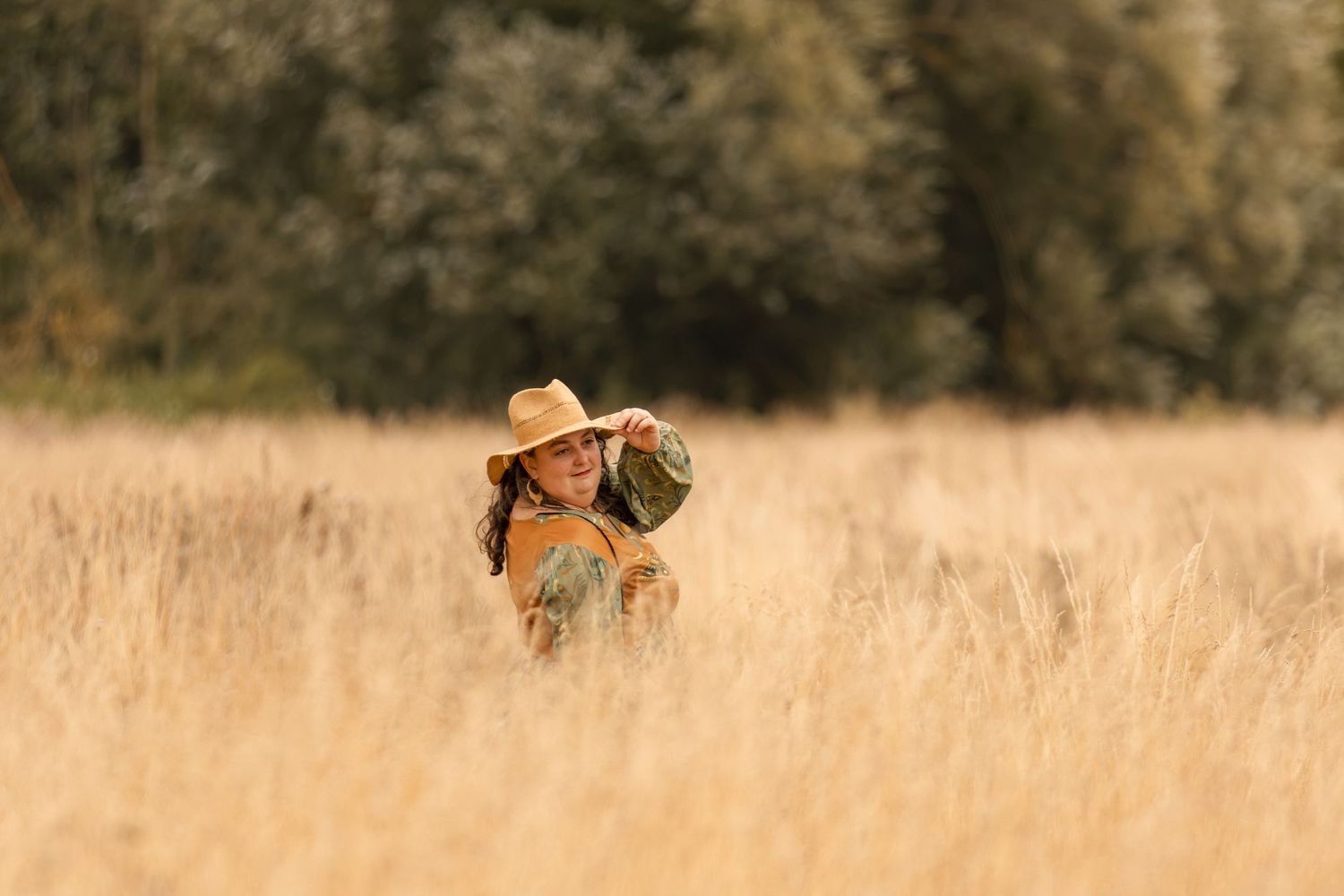 Woman in straw hat standing in golden wheat field with green trees in background on sunny day.