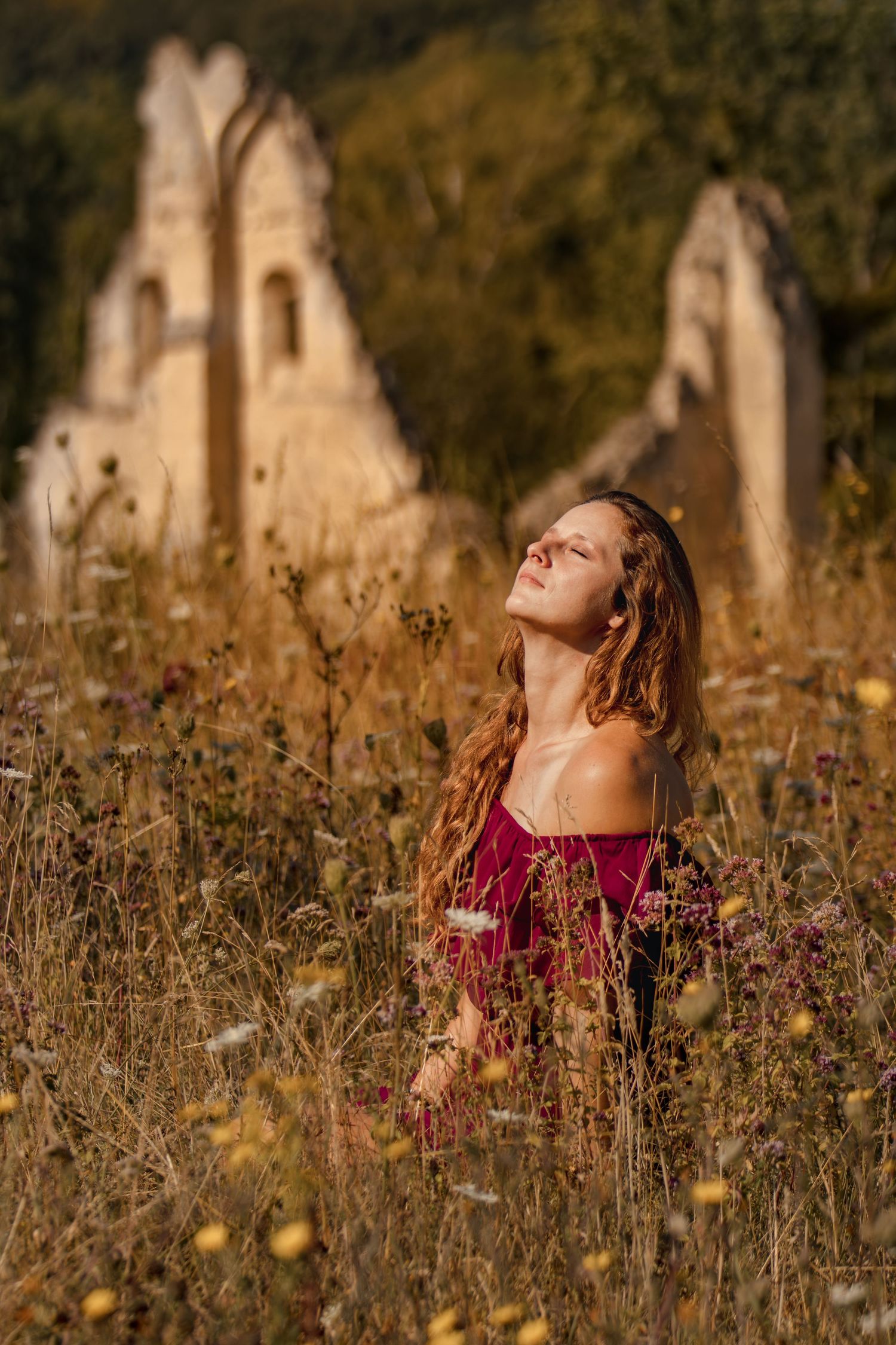 Young woman with long hair in red dress sits in wildflower field before ancient stone ruins at golden hour.