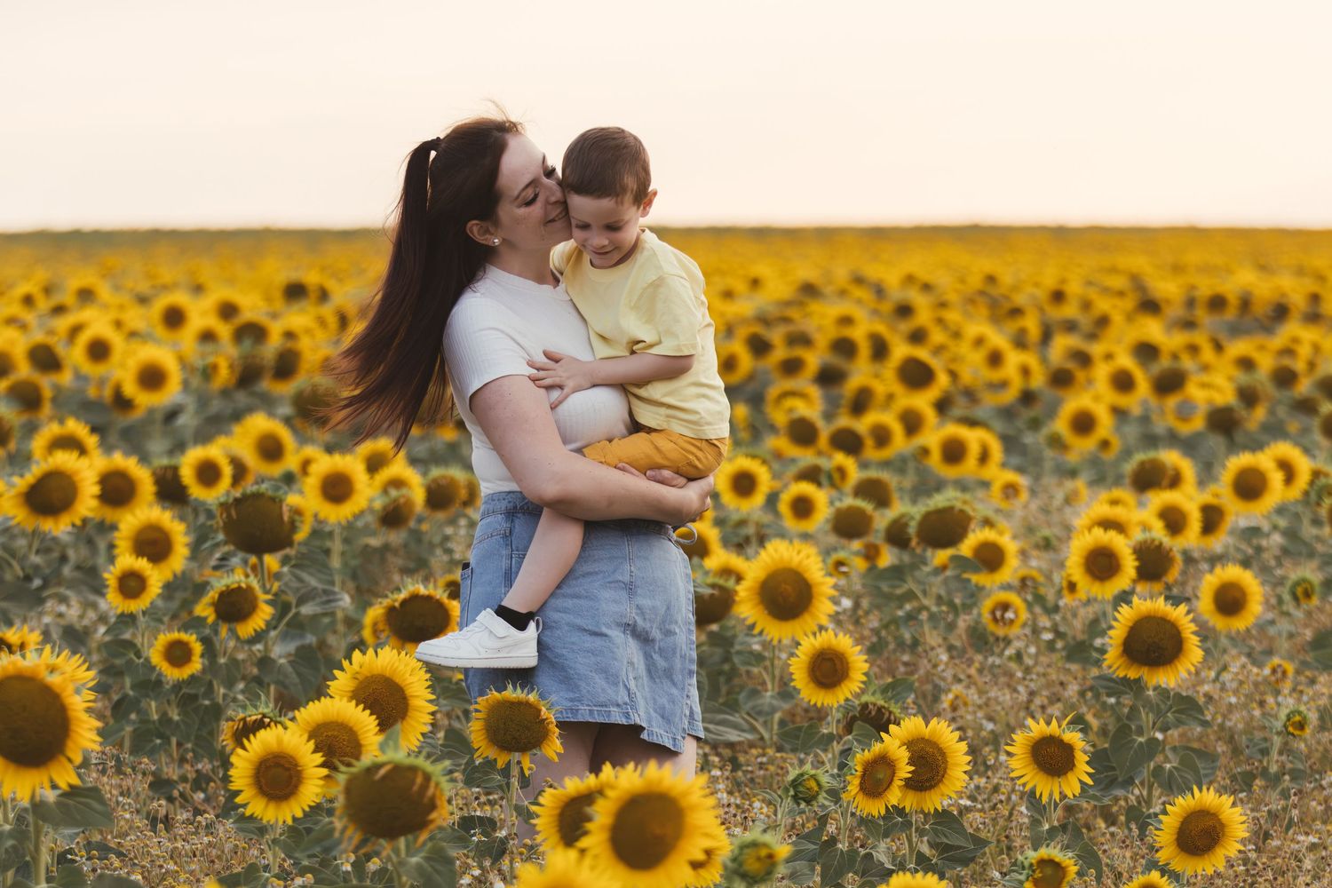 A mother holds her toddler in a vibrant sunflower field at sunset, both smiling joyfully.
