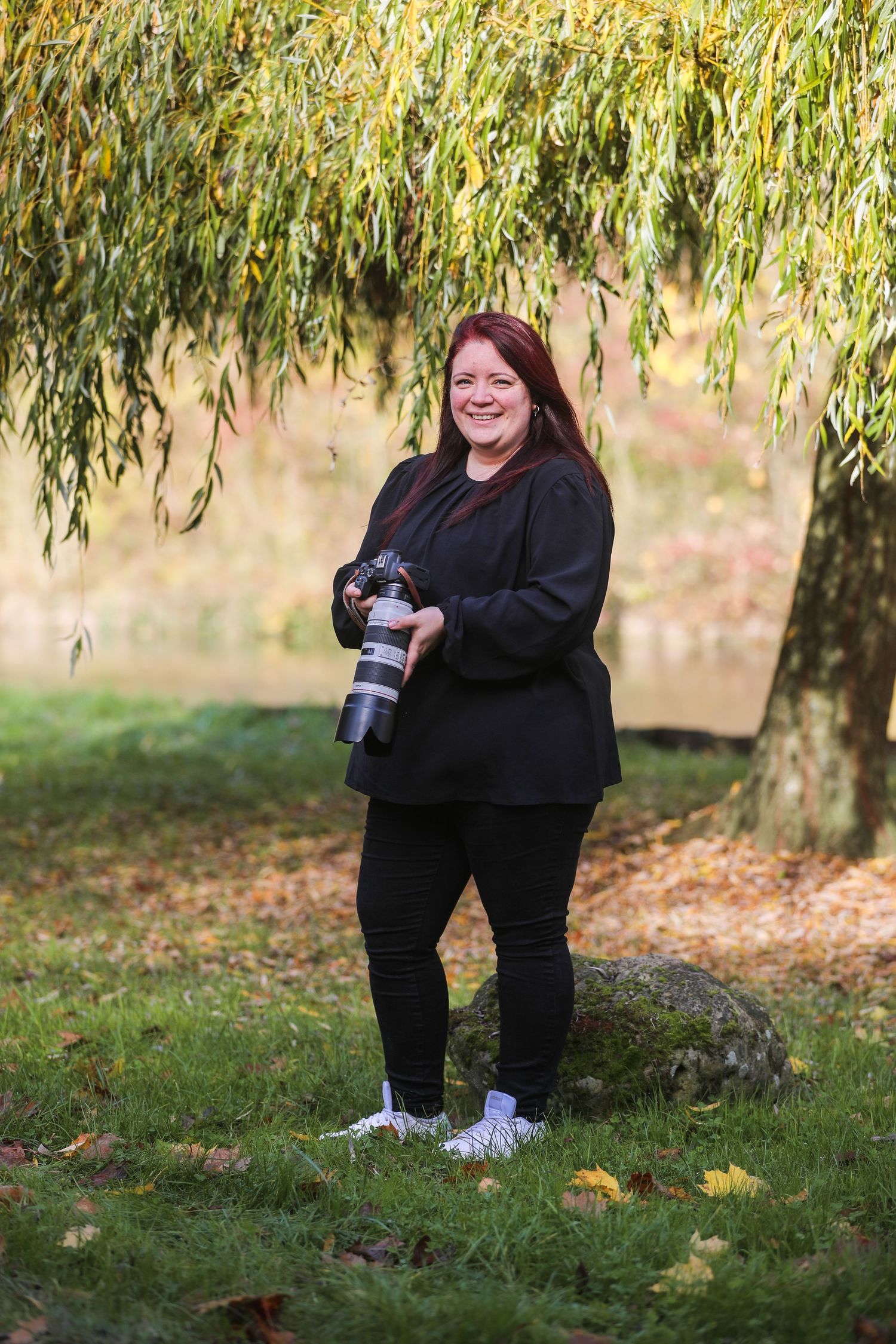 A smiling woman with red hair holds a professional camera under a weeping willow tree in autumn park.