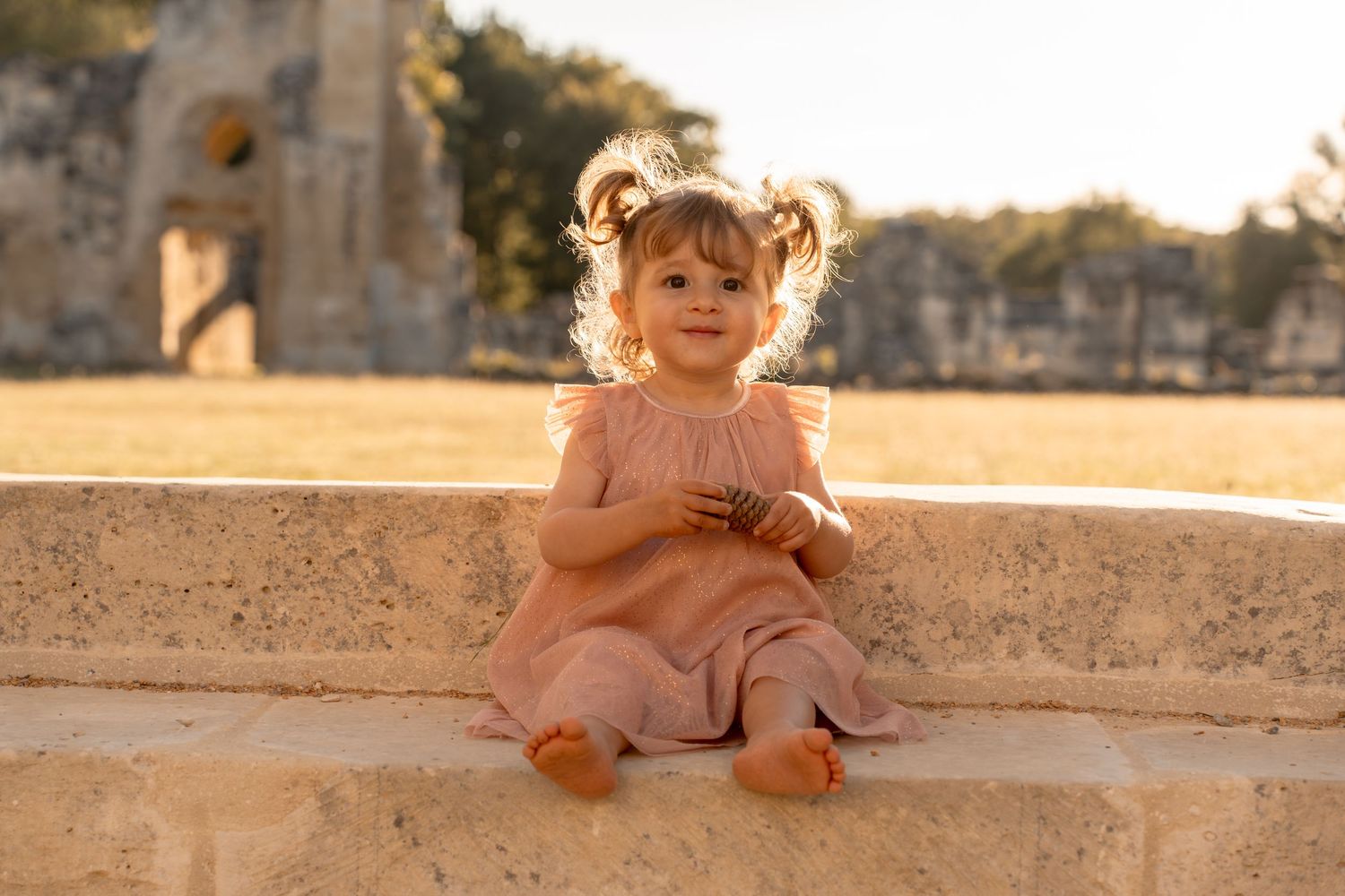 Adorable toddler girl in pink dress sits on stone steps at historic ruins during golden hour.