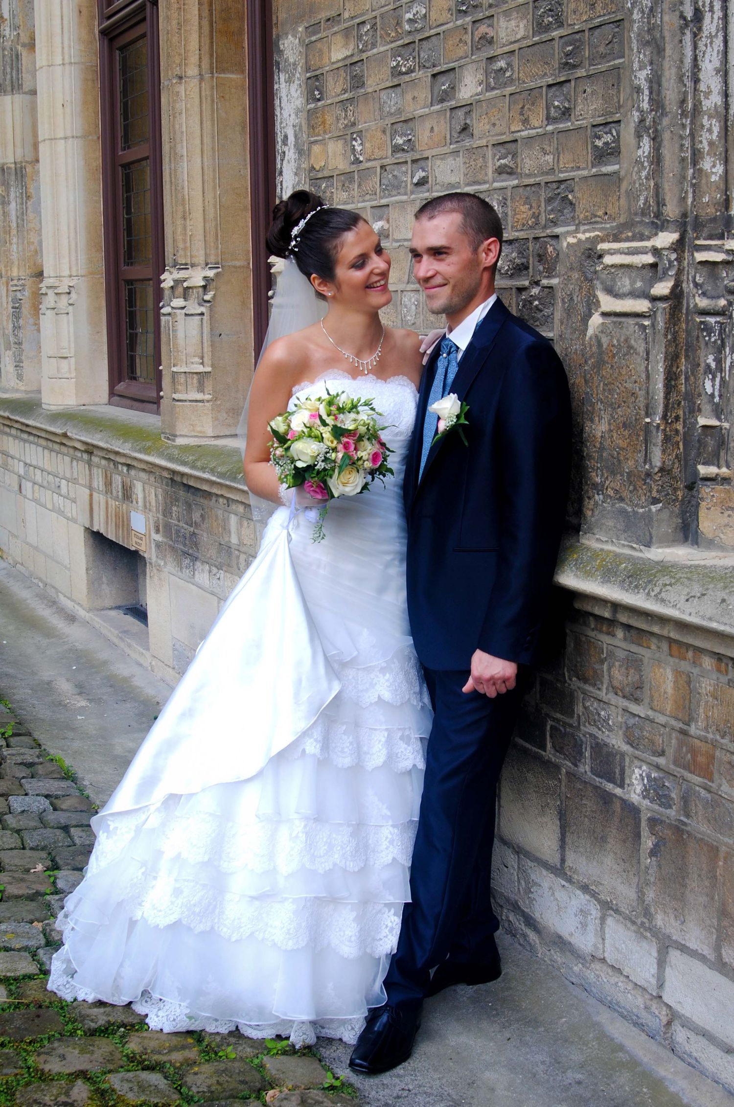 A smiling bride and groom posing together against a stone wall holding a pink and white bouquet.