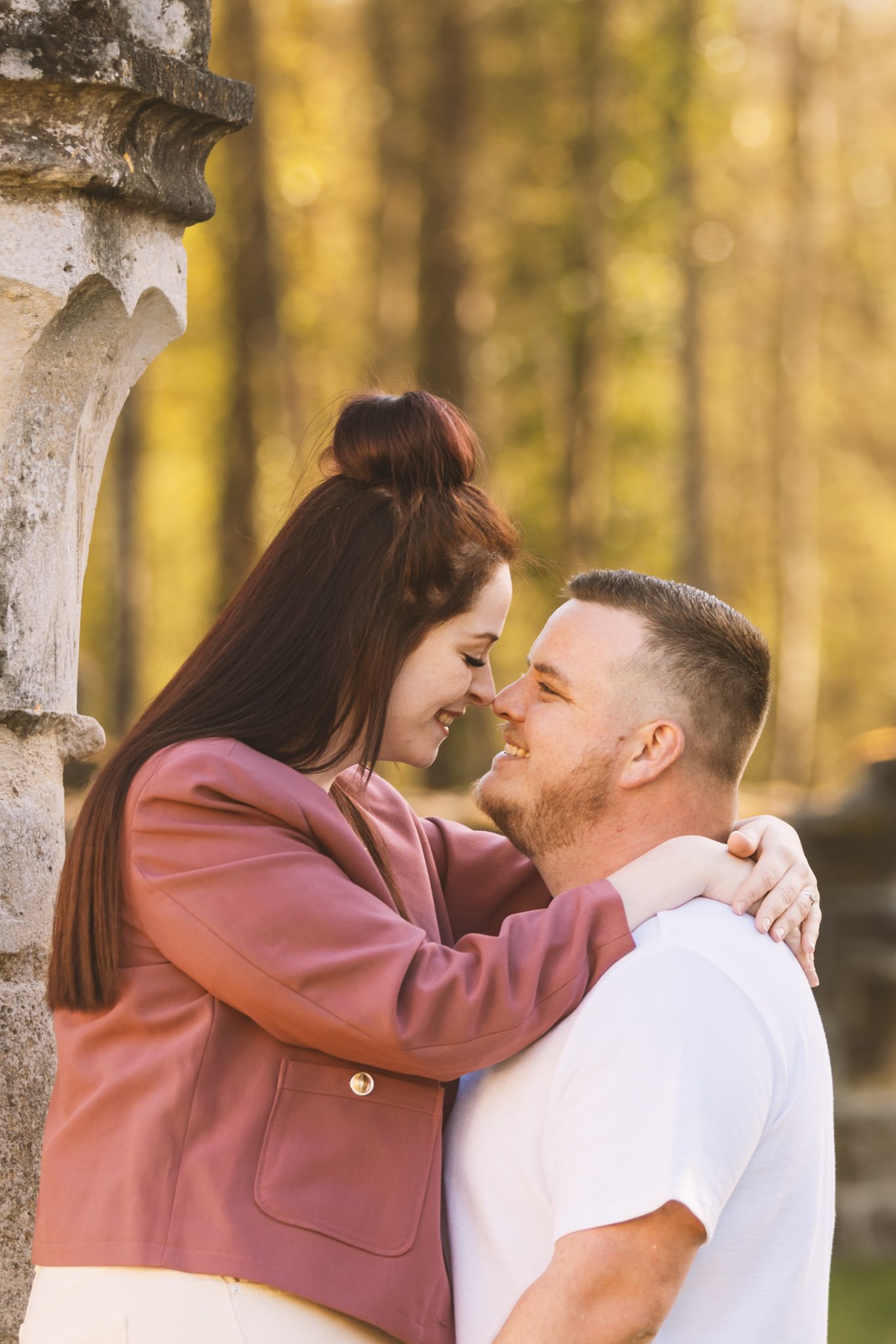 A couple shares a romantic moment outdoors, smiling at each other near a stone pillar with autumn trees in the background.