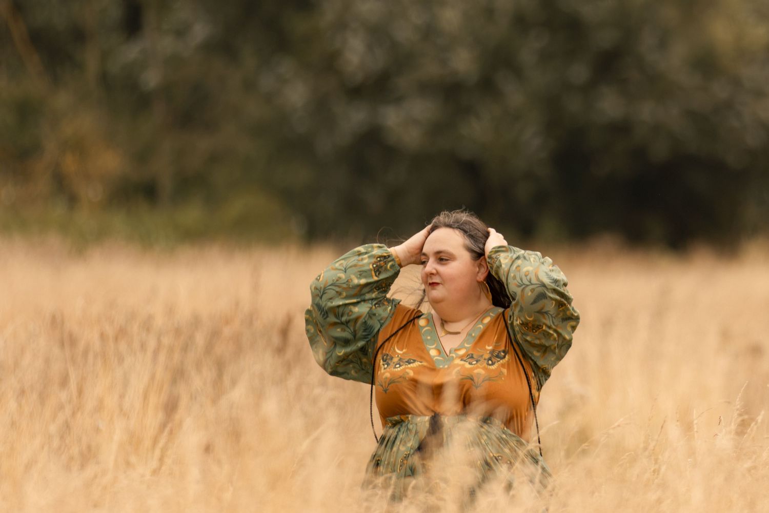 Woman in camouflage jacket standing in golden field, shielding eyes from sun with trees in background.