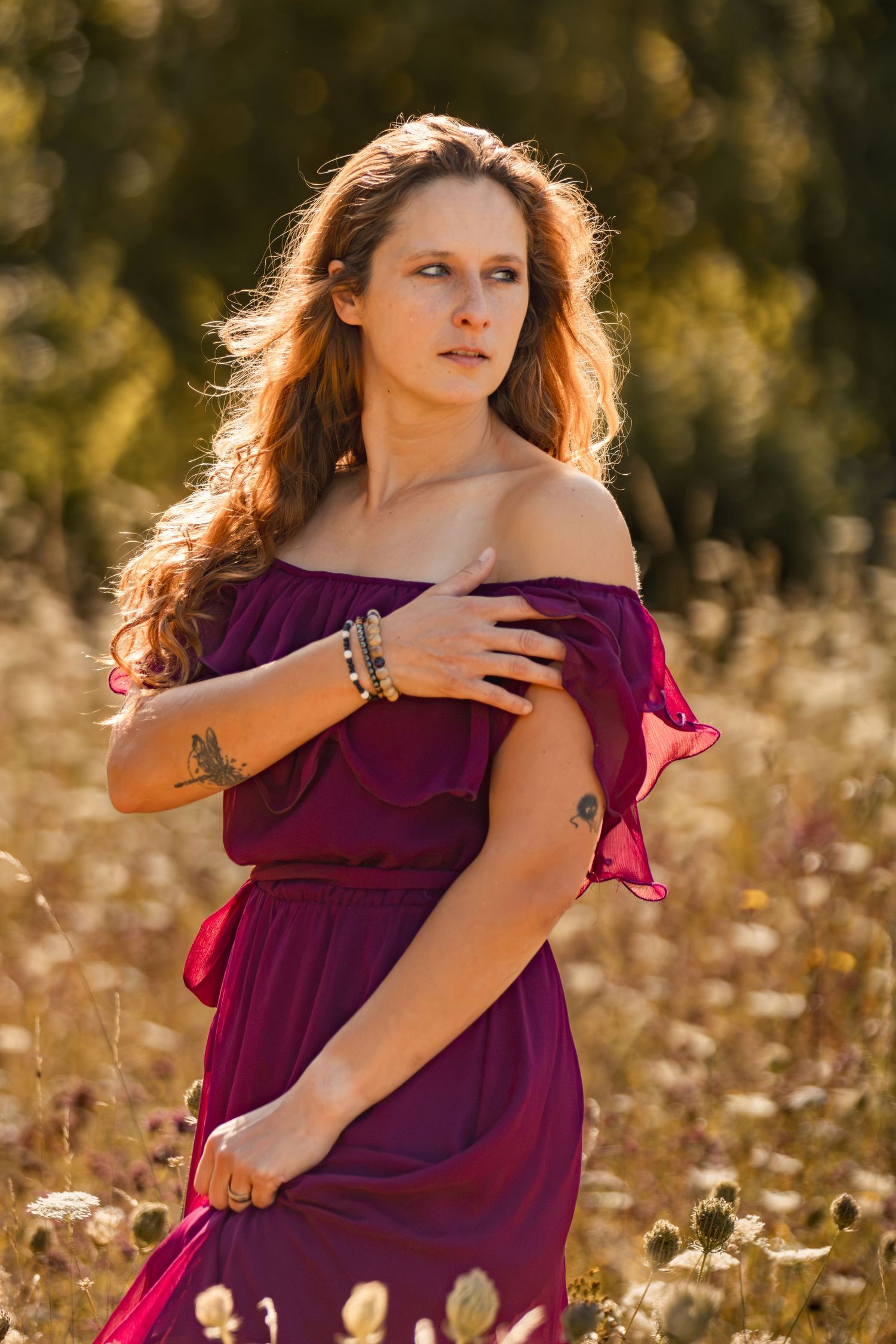 Woman in purple off-shoulder dress standing in golden field, long auburn hair, tattoo on wrist, bracelets.