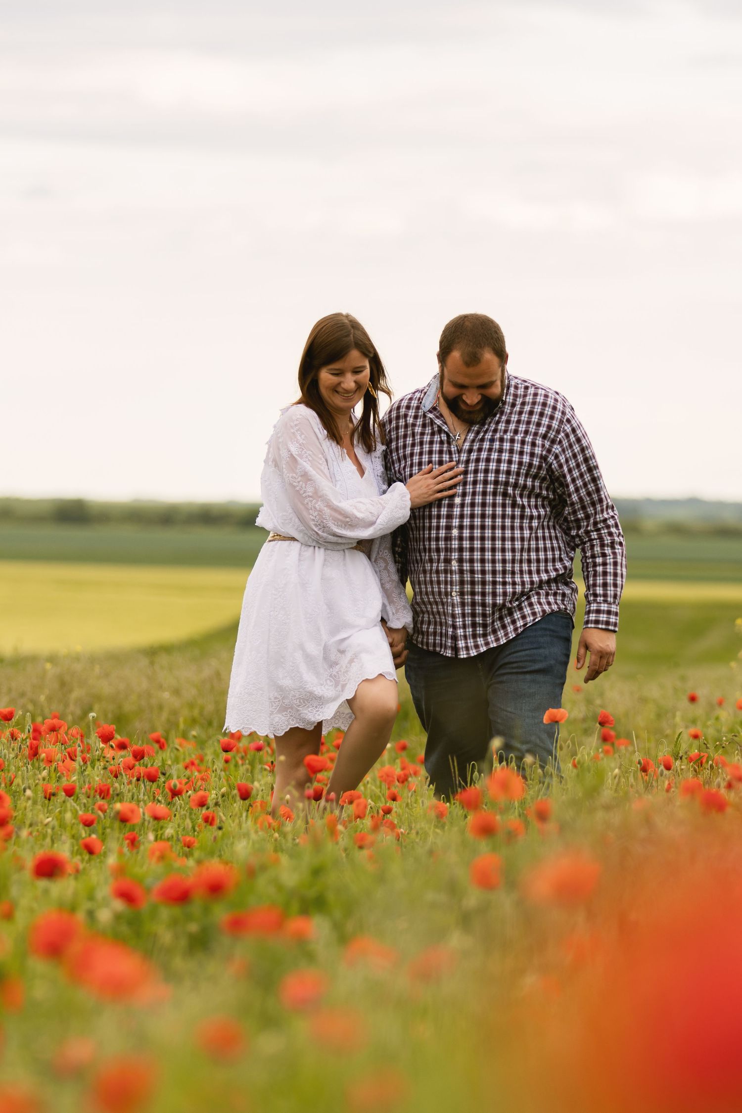 Couple walking through a vibrant red poppy field, woman in white dress with arm around mans shoulder.