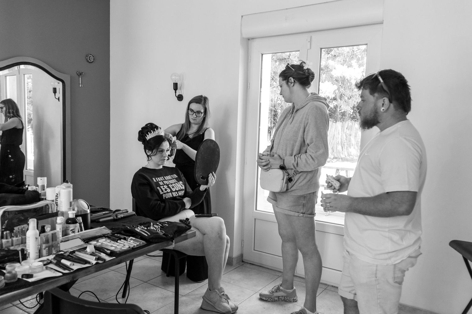 Black and white photo of a woman getting makeup done by a stylist while two people watch in a bright room.