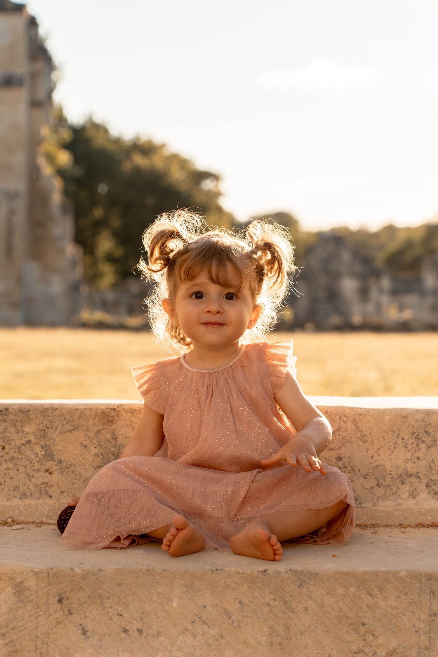 An adorable toddler girl in a pink dress sits cross-legged on a stone ledge at golden hour.