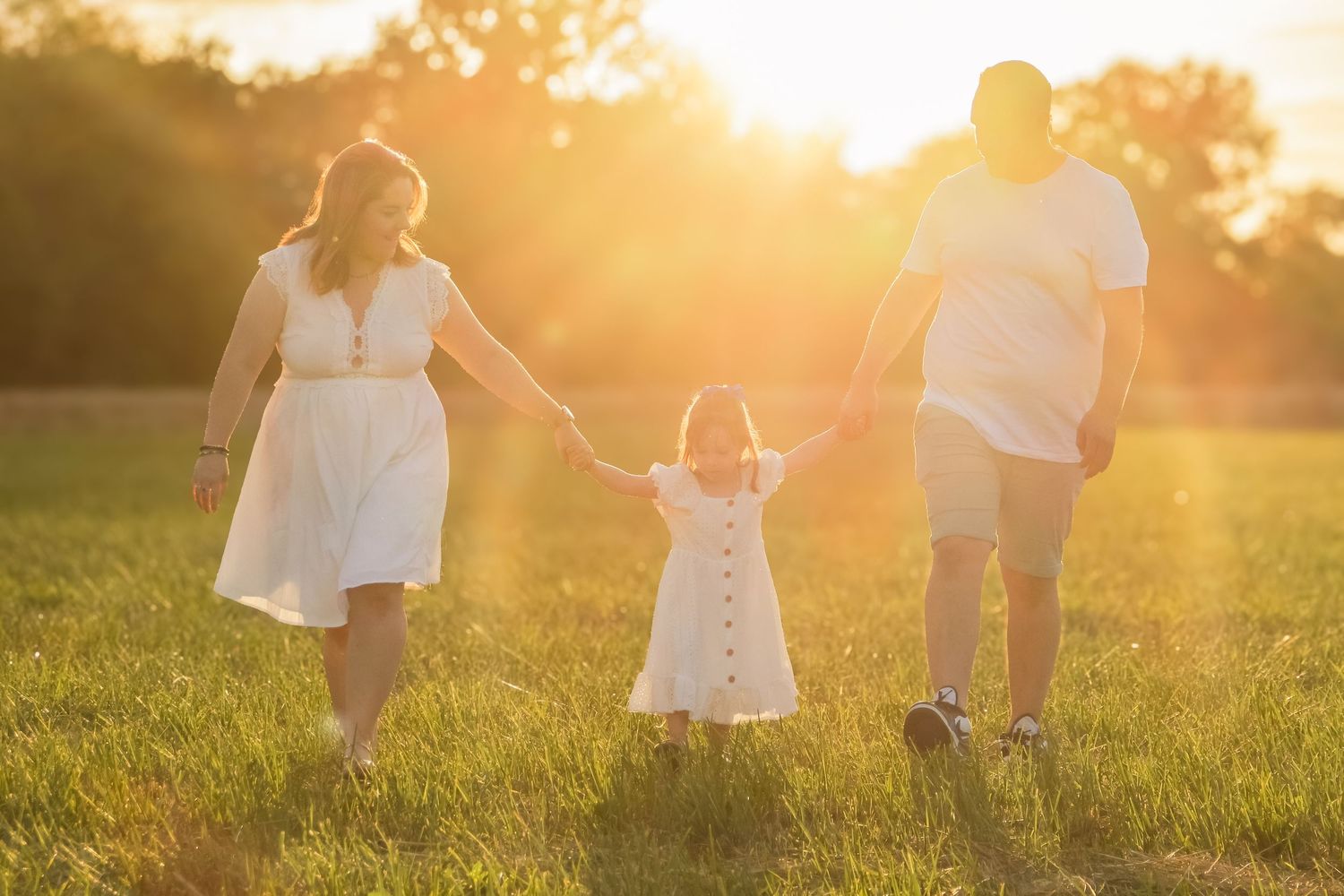 Family of three holding hands walking through a sunny green field at golden hour in matching white outfits.