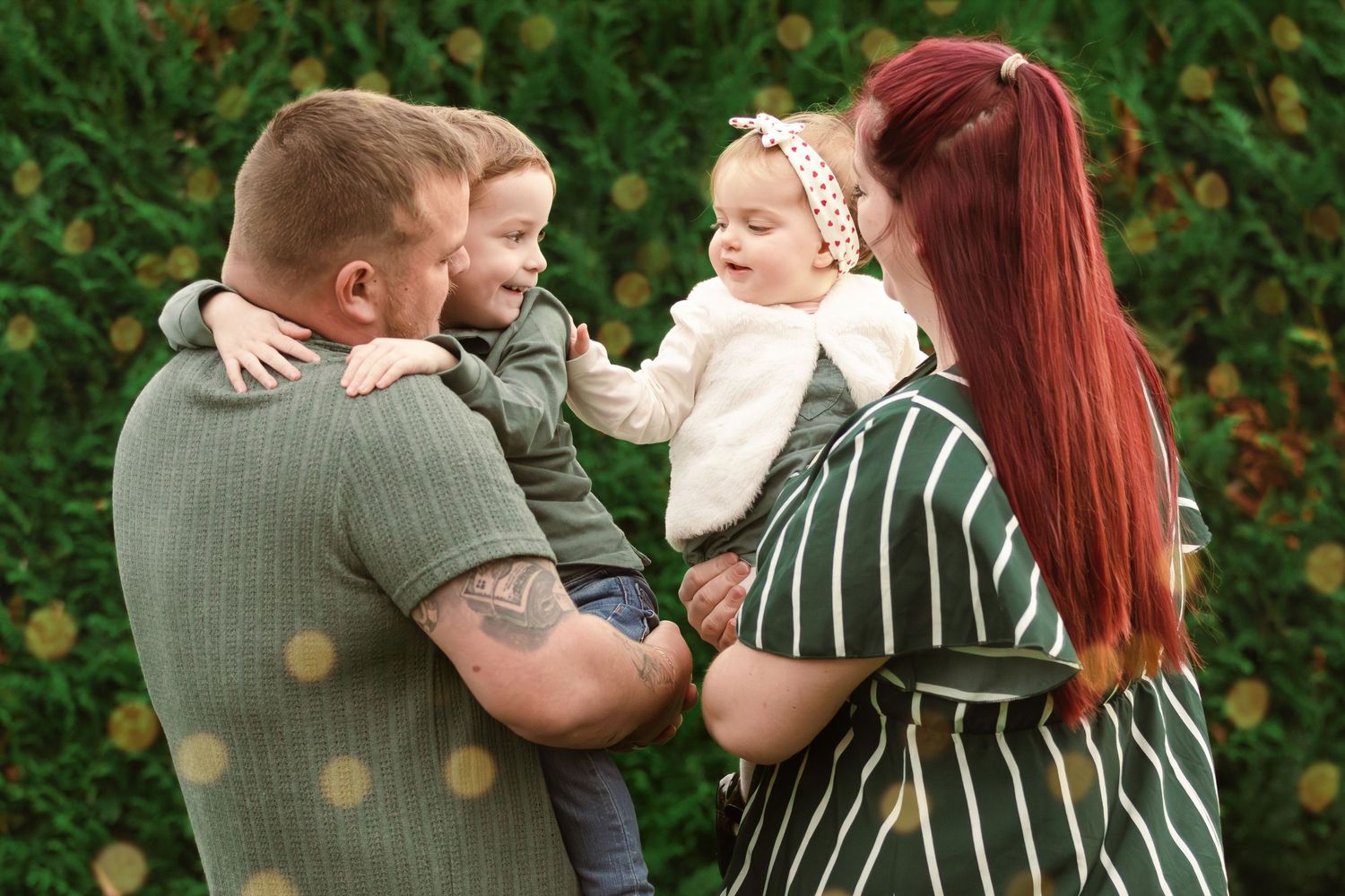 Parents holding their two young children share a joyful moment in a lush green garden.