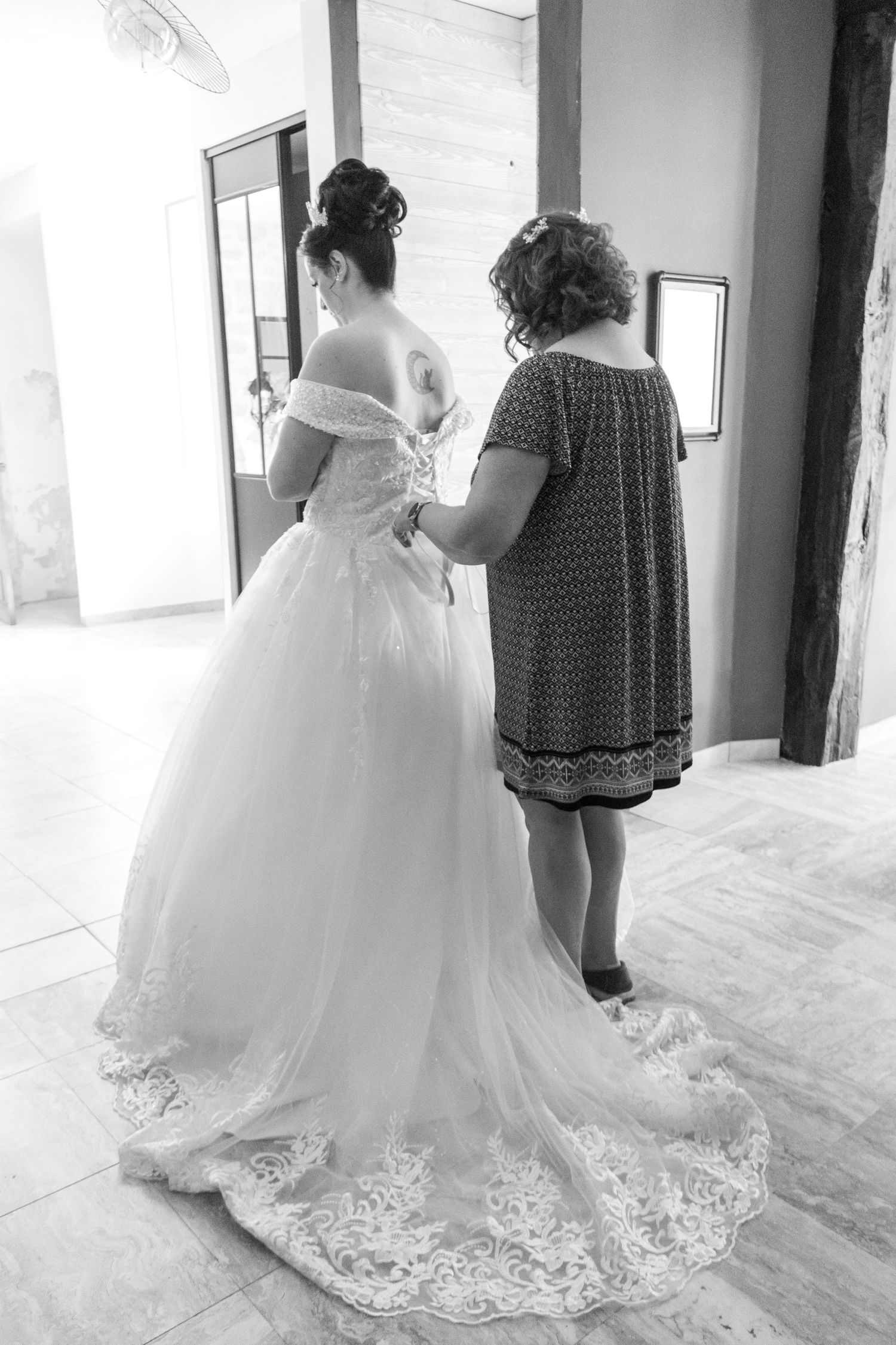 Black and white photo of a bride in a lace wedding gown being helped by a woman fastening the back of her dress.