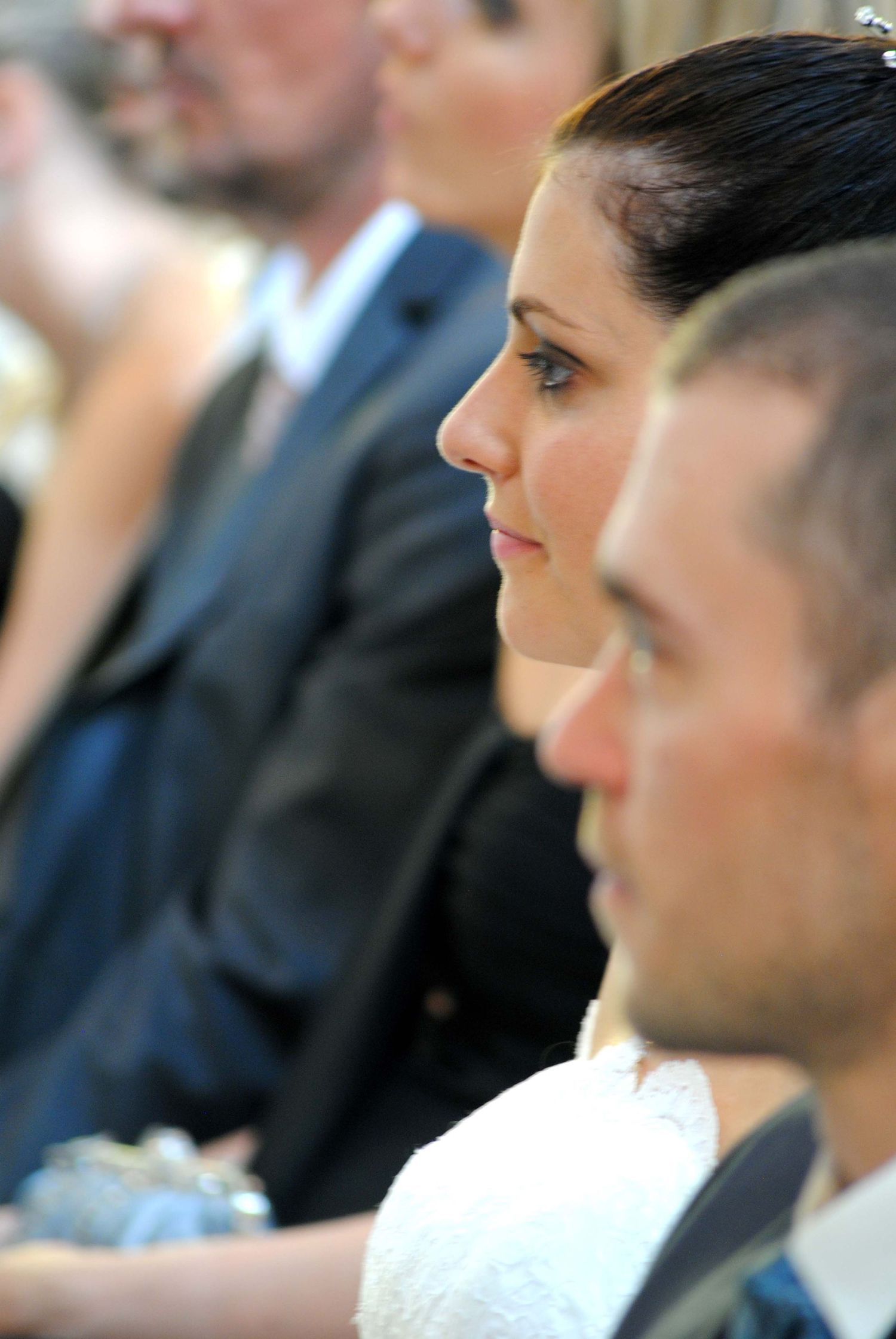 A bride in profile with elegant updo hairstyle attentively listening during a wedding ceremony.