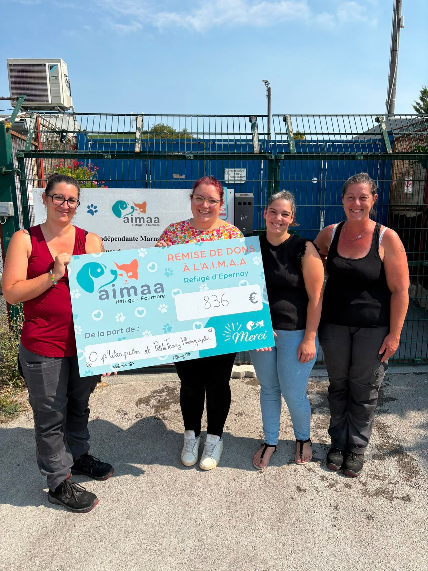 Four women holding a large donation check for AIMAA animal shelter outdoors in sunny weather.