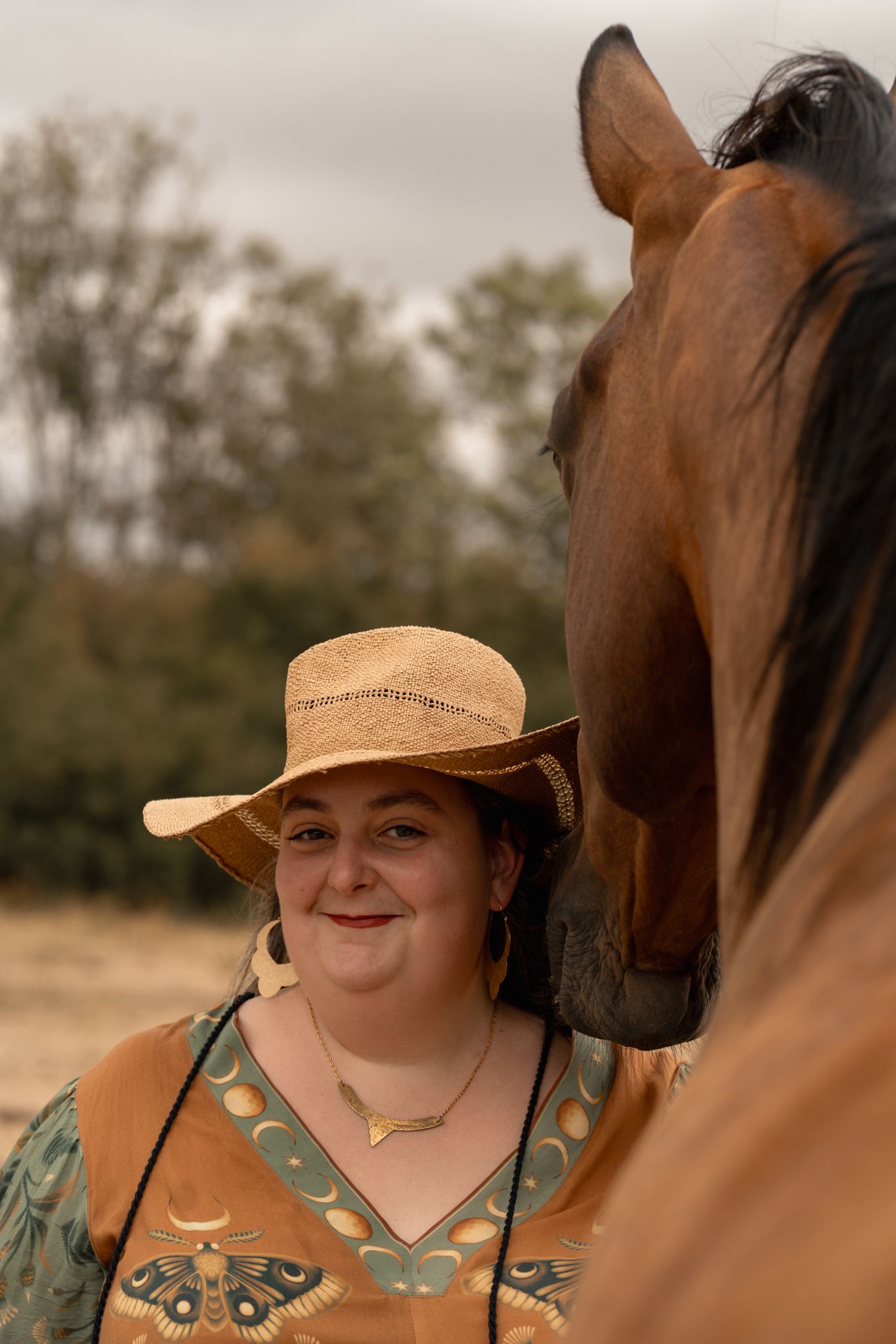 Woman in straw cowboy hat smiling beside a bay horse outdoors on a cloudy day.
