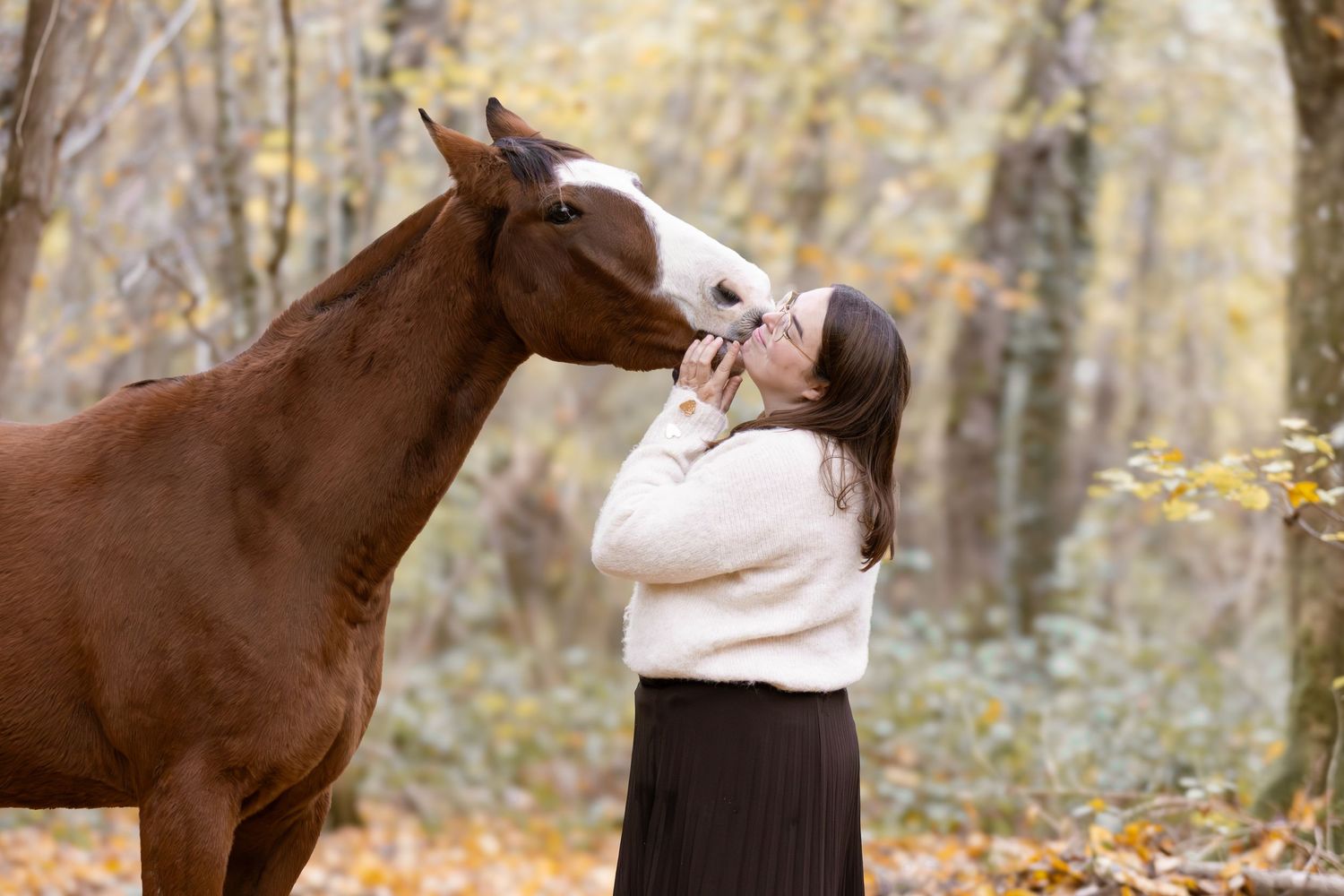Woman in white sweater kissing a brown horse with white blaze in fall foliage setting.