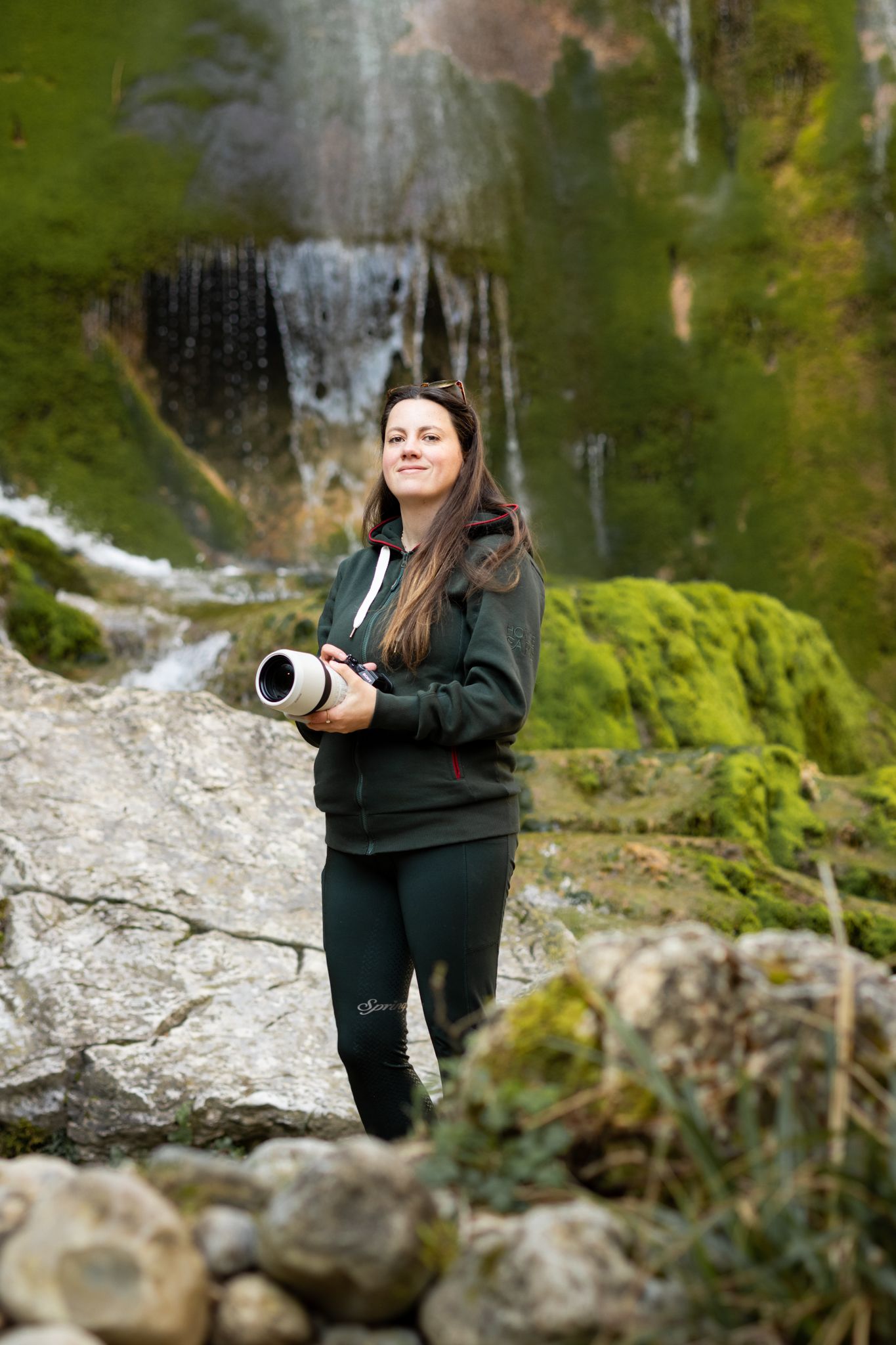 Woman in green tracksuit holding camera lens stands before a mossy waterfall in a lush forest setting.