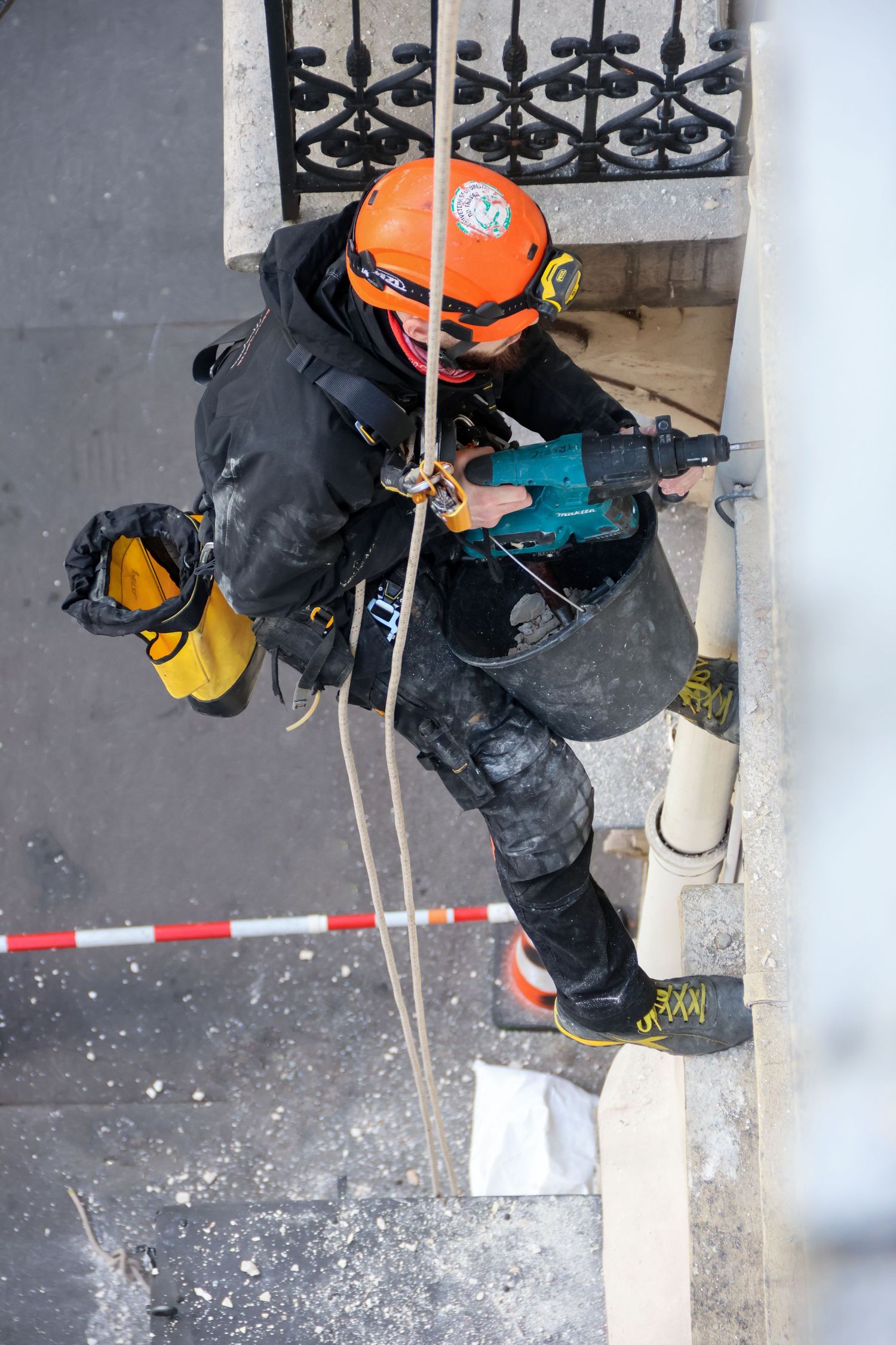 Worker in orange helmet rappelling down building facade using power drill for maintenance work.