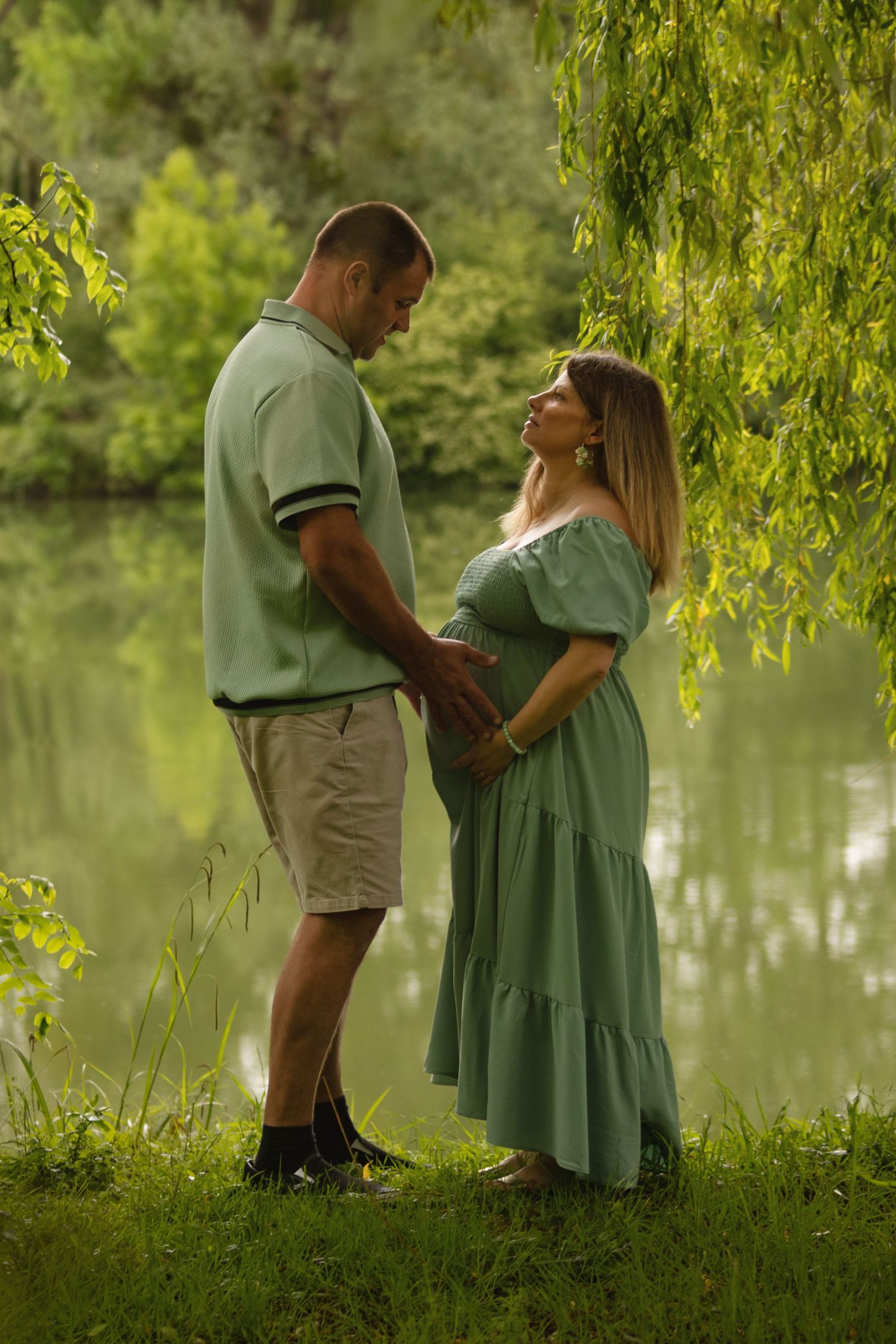 Couple holding hands by a serene pond, woman in green maternity dress, man in mint polo, surrounded by lush greenery.