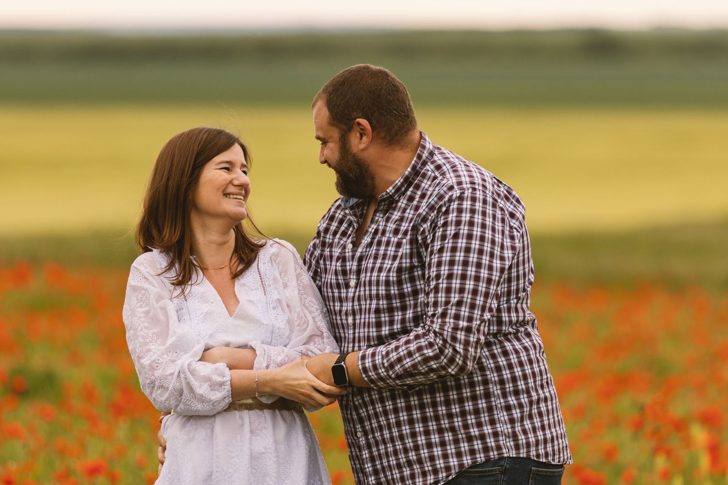 Couple smiling at each other in a vibrant red poppy field, woman in white dress, man in plaid shirt.