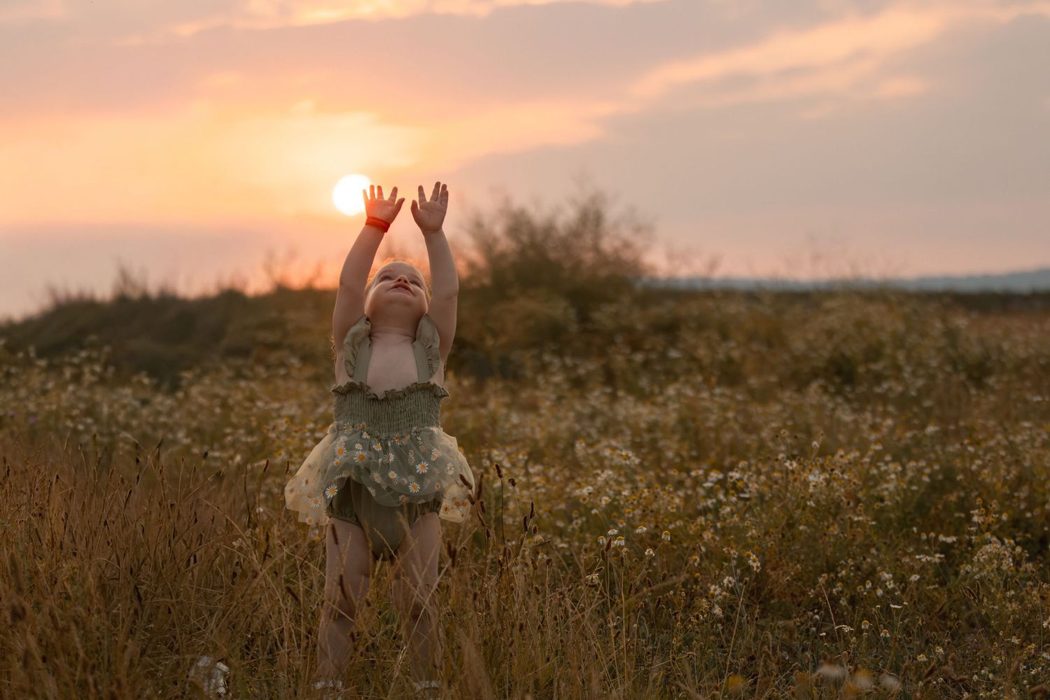 Child in floral dress raises arms toward golden sunset sun in wildflower meadow at dusk.