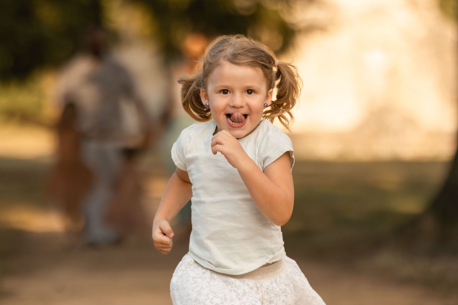 A joyful young girl with pigtails runs outdoors in a sunny park, laughing and wearing a white top and skirt.