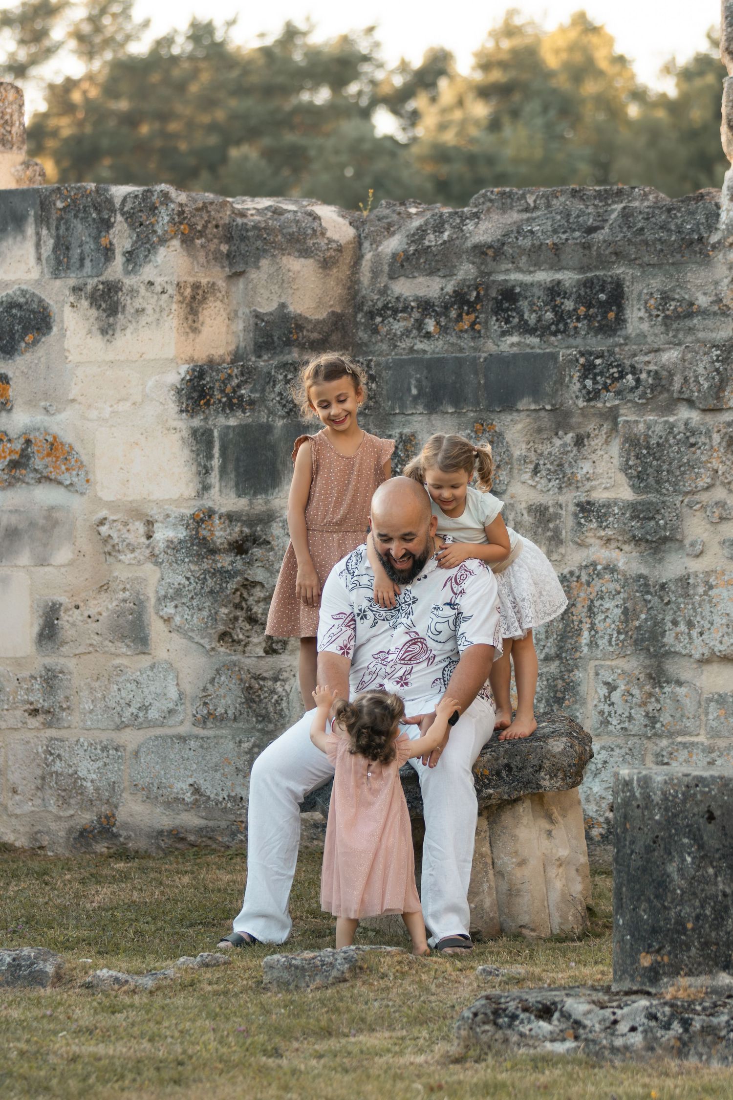 Bald father sits on stone ruins laughing with three young daughters in pink and white dresses outdoors.