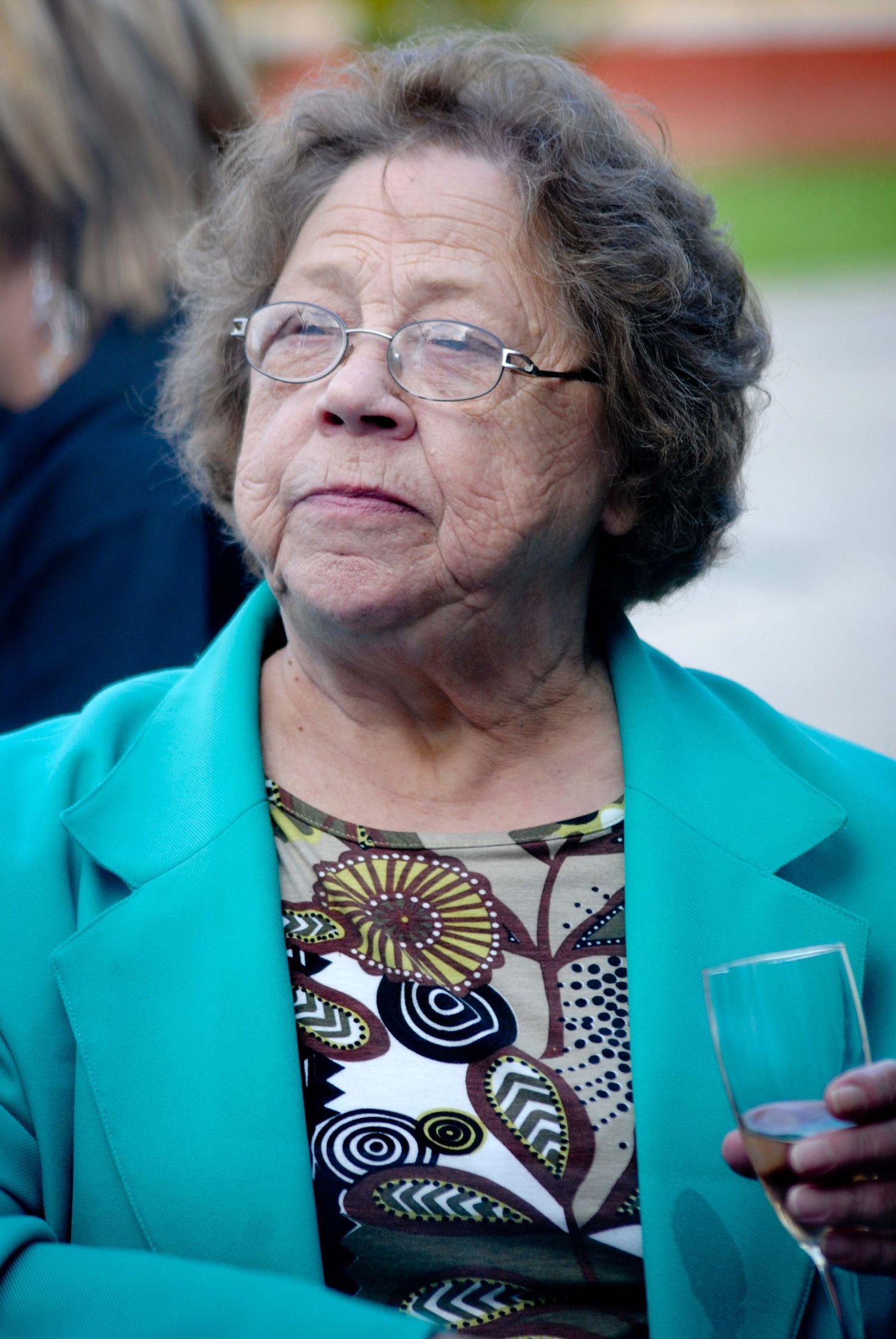 Elderly woman wearing teal blazer and floral blouse, glasses, holding a glass at an outdoor event.
