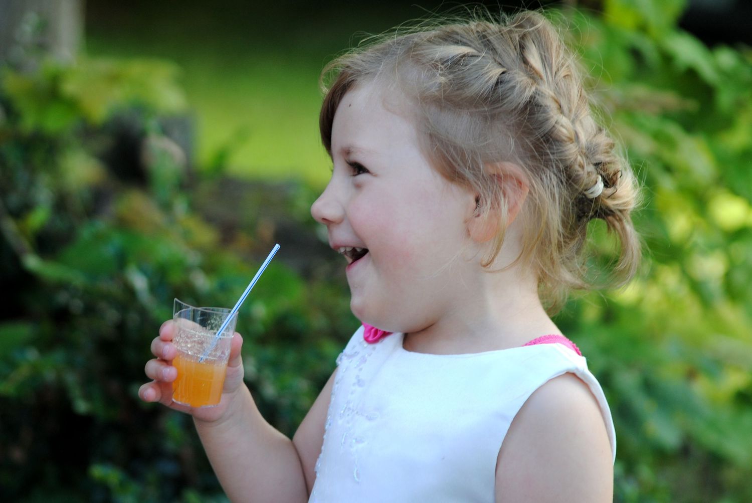 A happy toddler girl with blonde hair holds a glass of orange juice with a blue straw outdoors.