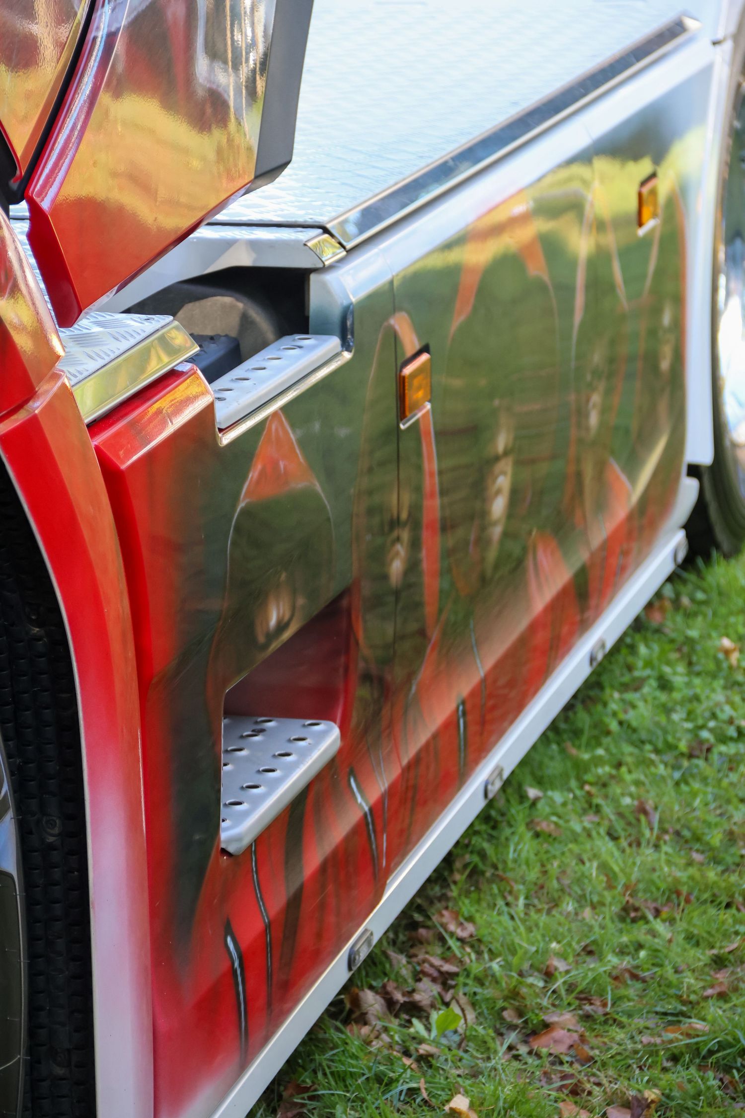 Shiny red and chrome custom truck side panel reflecting green trees in its polished metallic surface.