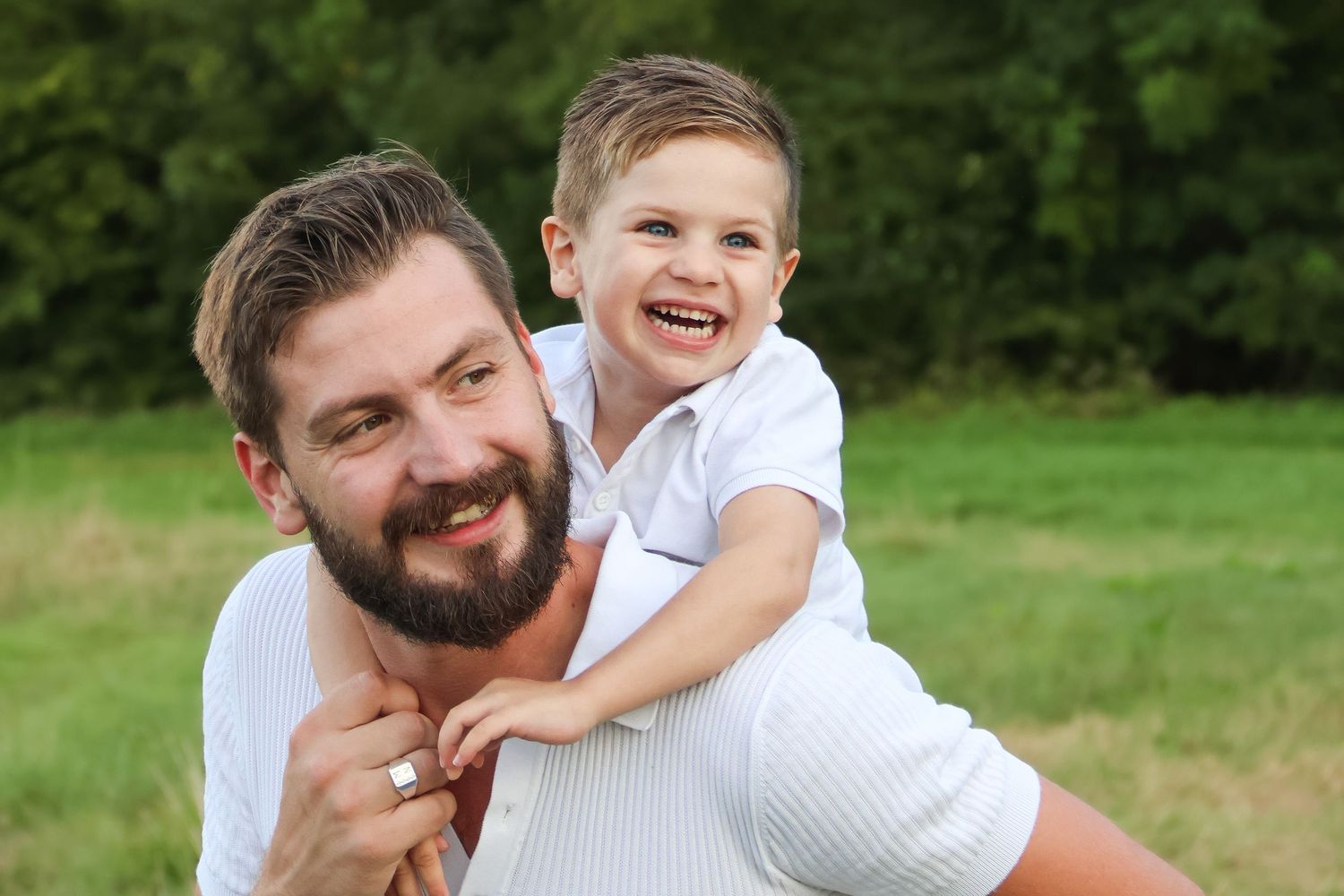 Bearded father giving his laughing young son a piggyback ride outdoors in a green park.