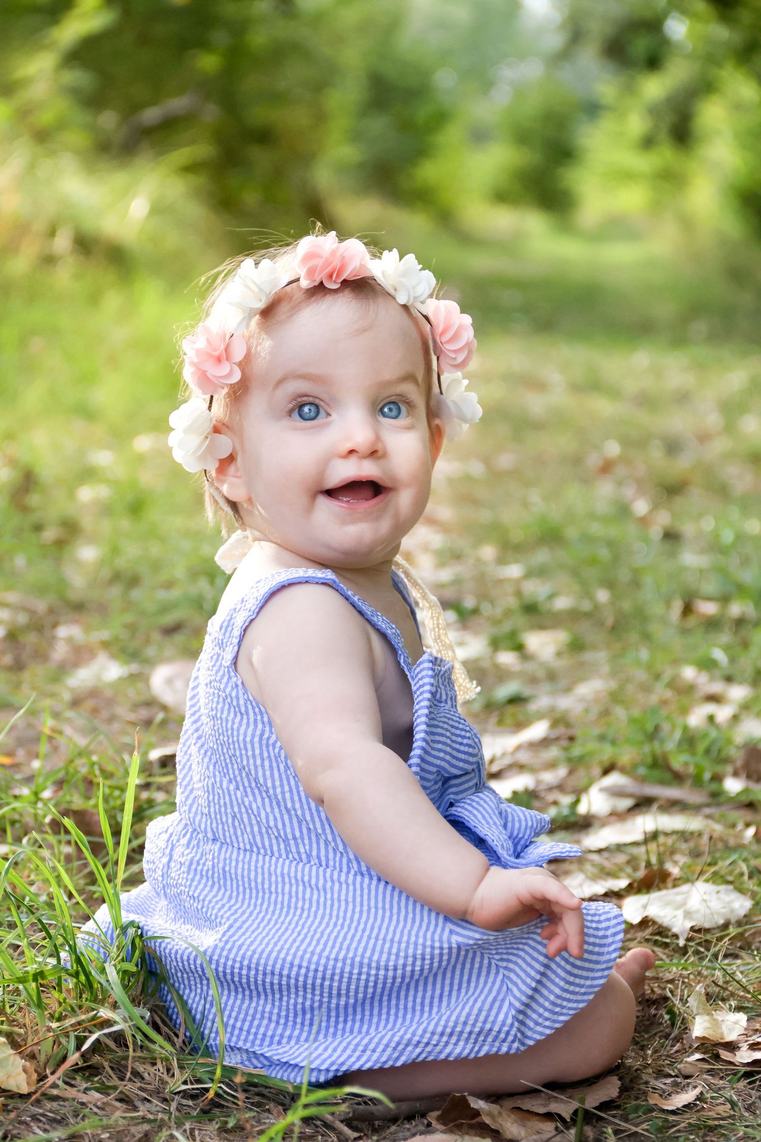 Adorable baby girl in blue striped dress and pink floral crown sitting on grass smiling outdoors.
