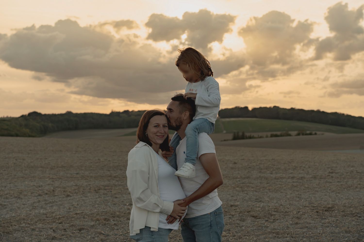 Family of three posing at sunset in an open field, father holding child on shoulders beside smiling mother.