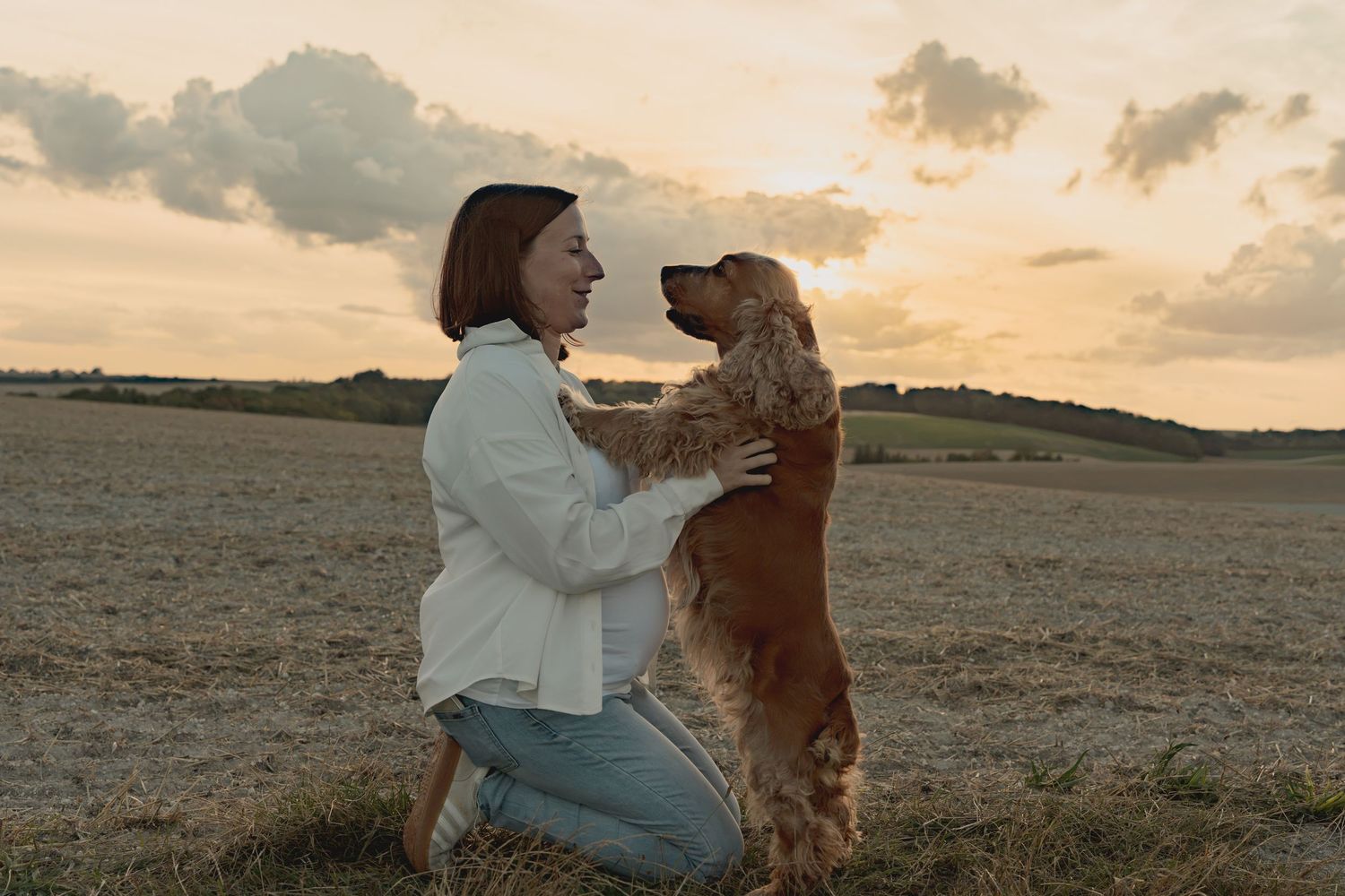 Woman kneeling in a field at sunset, lovingly holding a golden Cocker Spaniel standing on its hind legs.