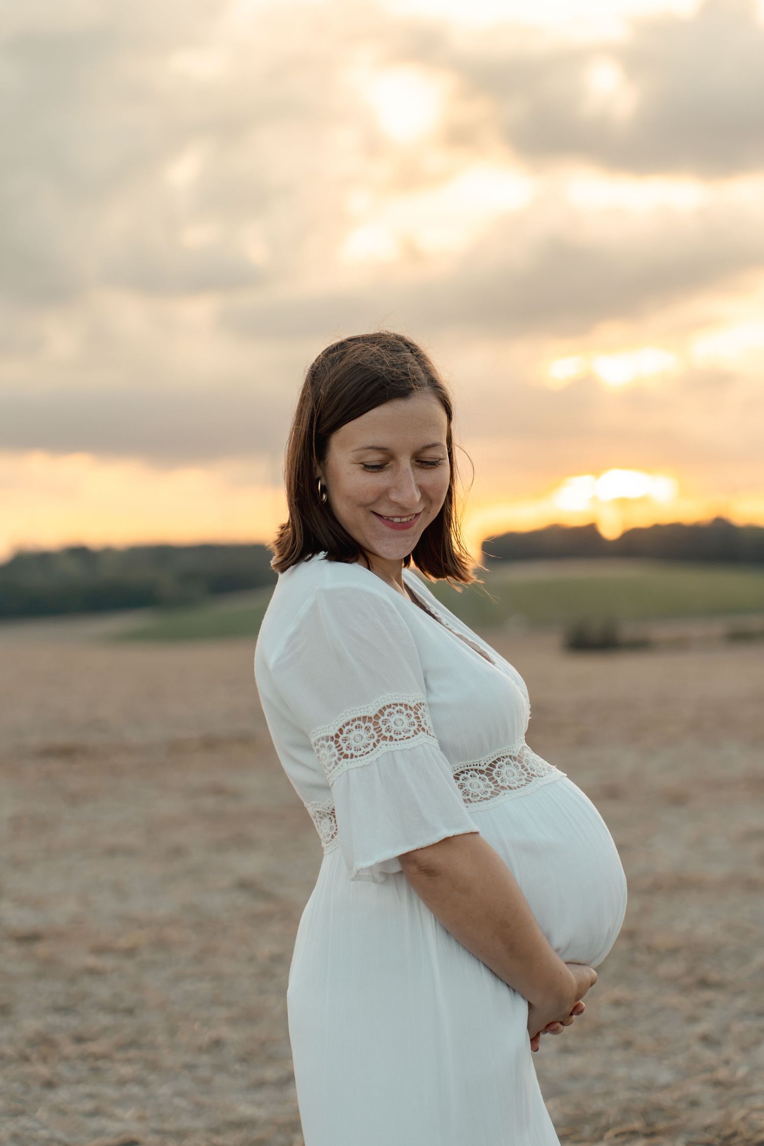 Pregnant woman in white dress cradling her baby bump at sunset in a golden field.