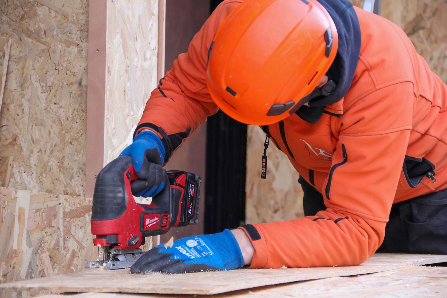 Worker in orange jacket and helmet uses Milwaukee jigsaw on wood panel at construction site.