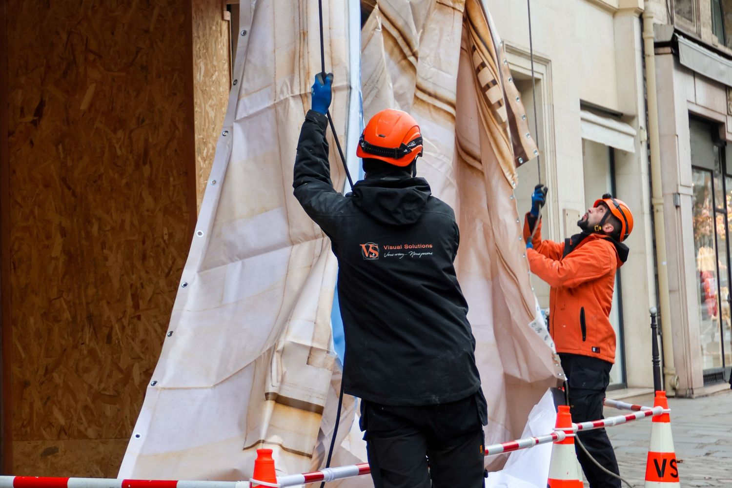 Two workers in orange helmets carefully installing protective covering on a building facade during renovation work.