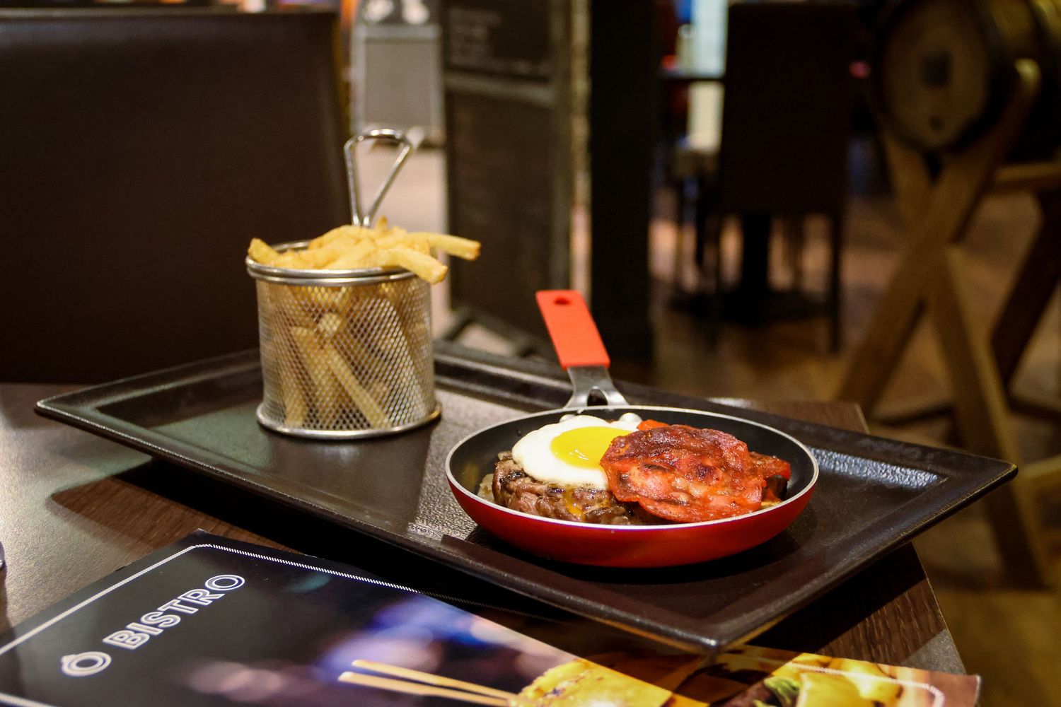 Sizzling skillet with meat, fried egg, and tomato sauce served alongside crispy french fries in a metal basket on a dark tray.